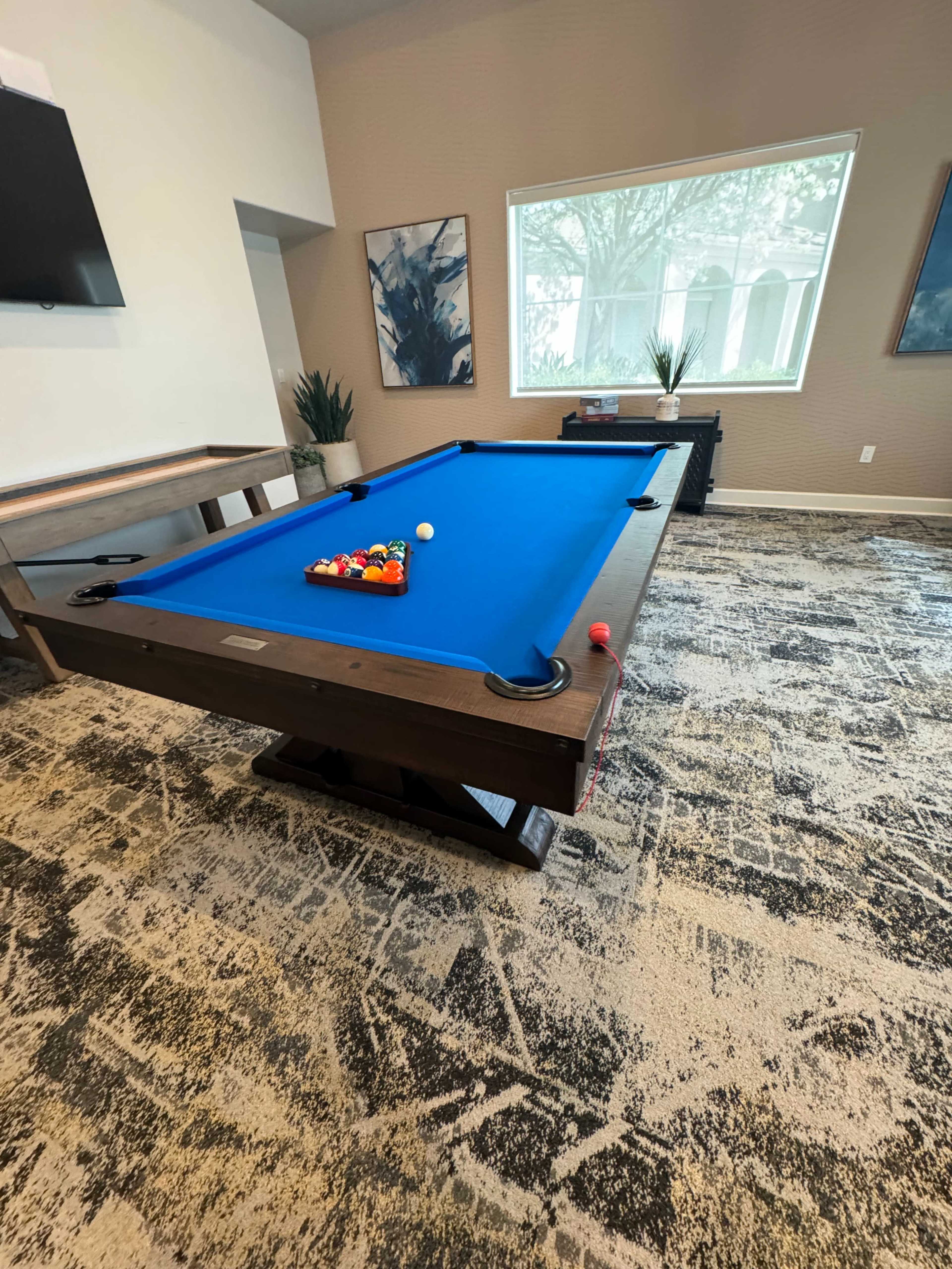 A blue pool table with a triangle of colored balls is set in a room featuring large windows and a patterned carpet.