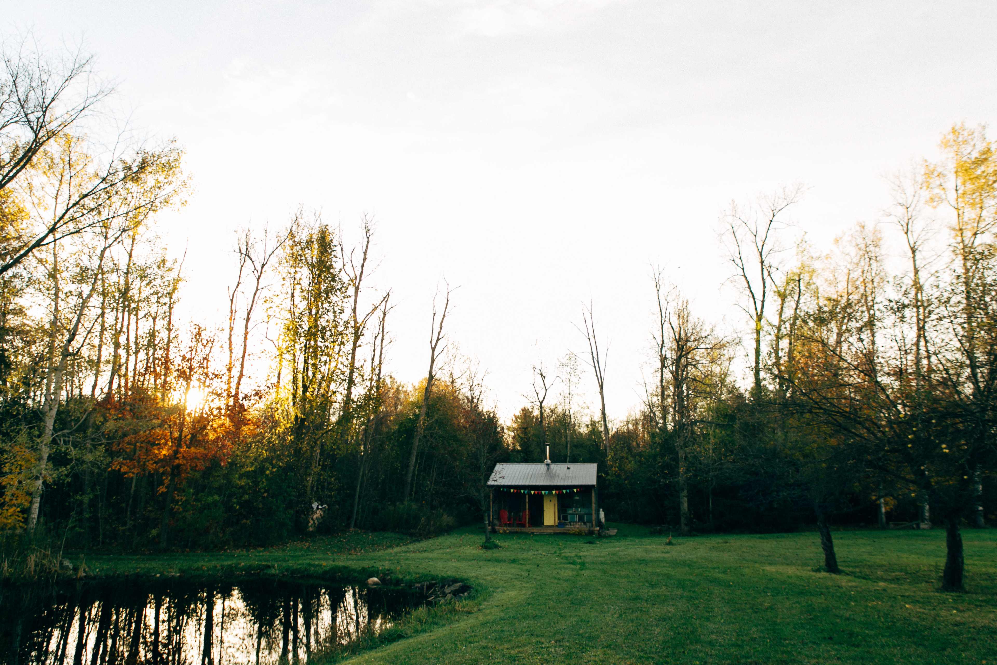 A small cabin with a metal roof is positioned near a pond, surrounded by trees as the sun sets in the background.