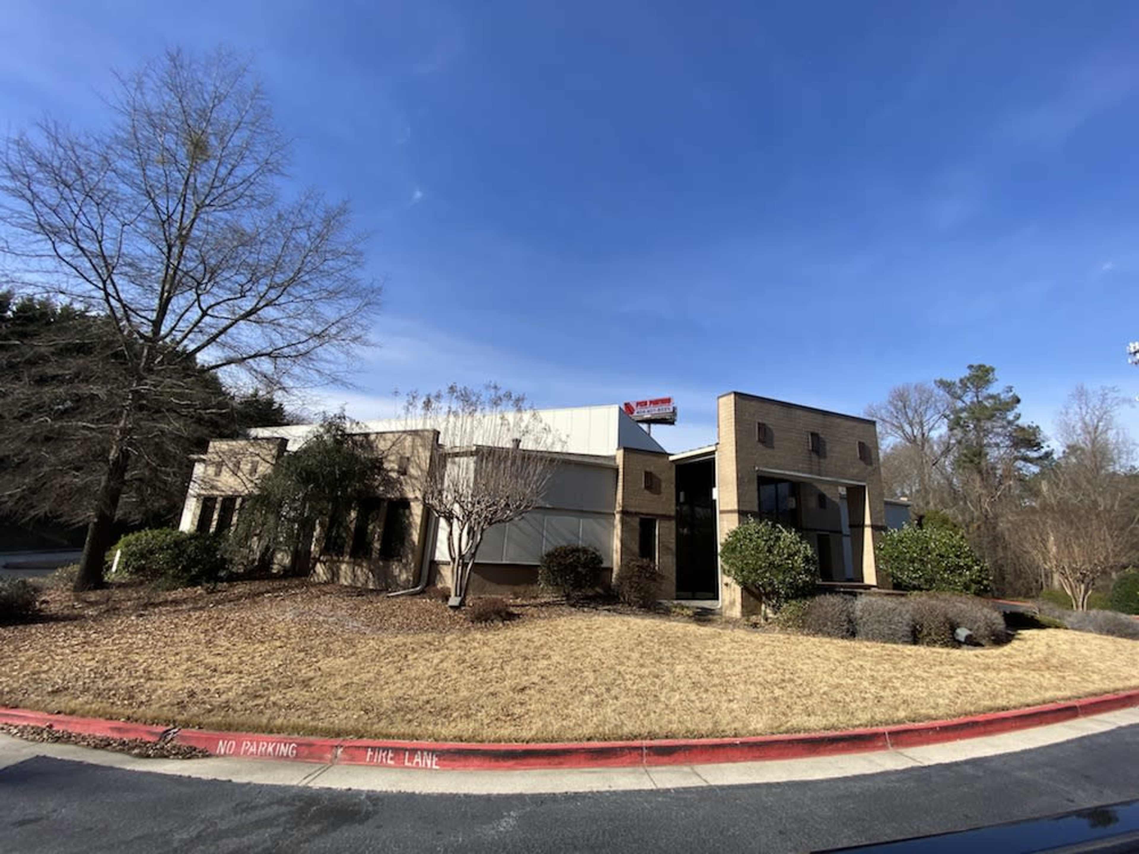 A two-story commercial building with a tan exterior and landscaped grounds is visible on a sunny day.