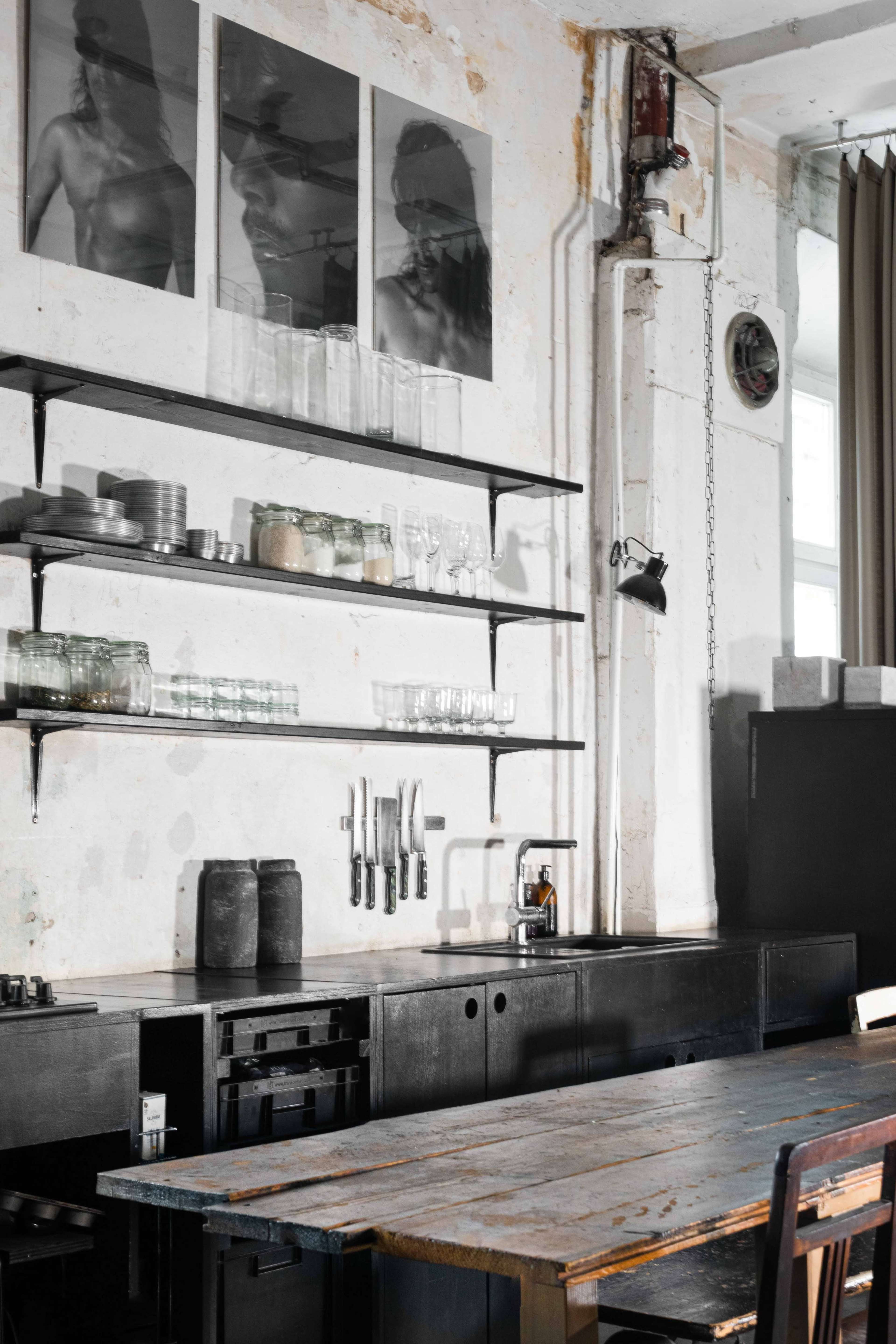 A minimalist kitchen with black shelves holding glassware and utensils, a wooden table, and black cabinetry along a textured white wall, featuring three framed black-and-white photographs.
