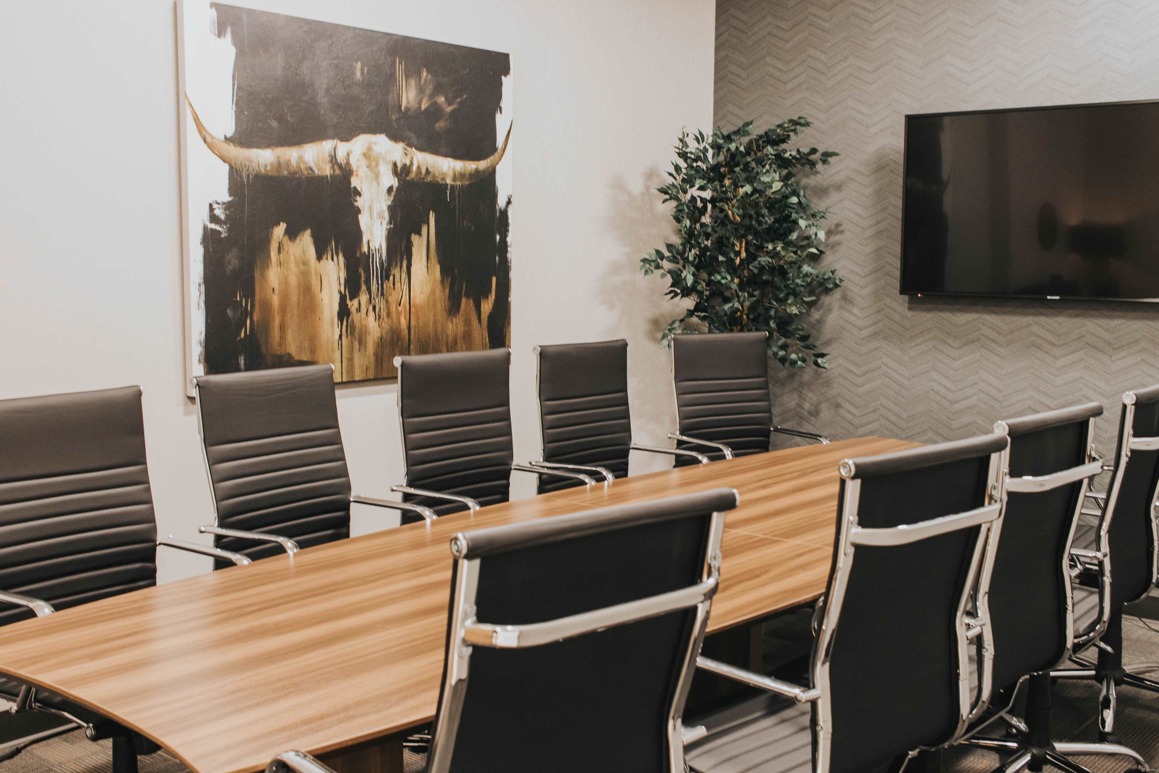 A conference room with a long wooden table, black ergonomic chairs, a large wall art featuring a bull's skull, and a flat-screen television mounted on the wall.