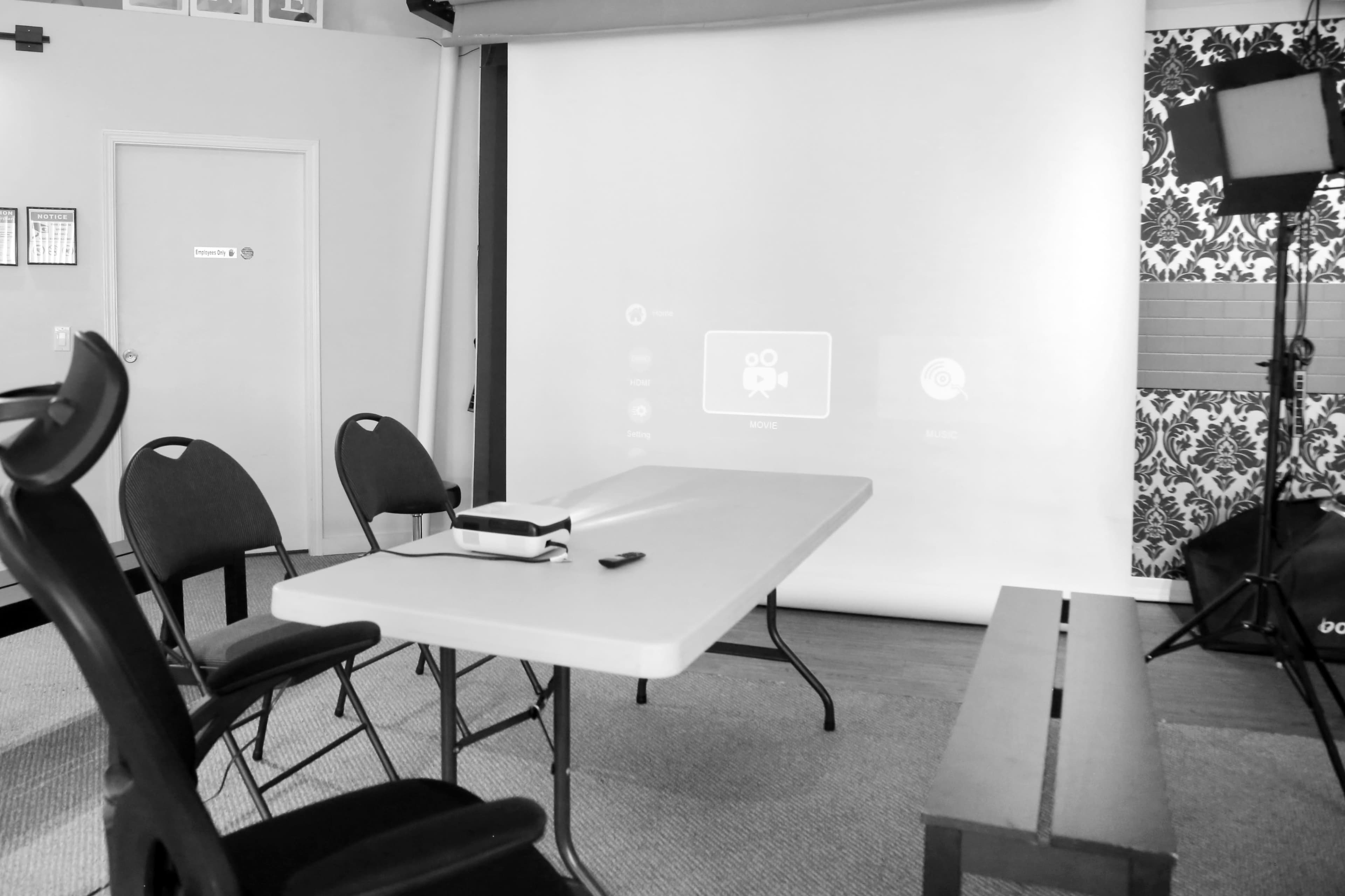 A table with a projector screen behind it, surrounded by chairs in a room with patterned wallpaper.