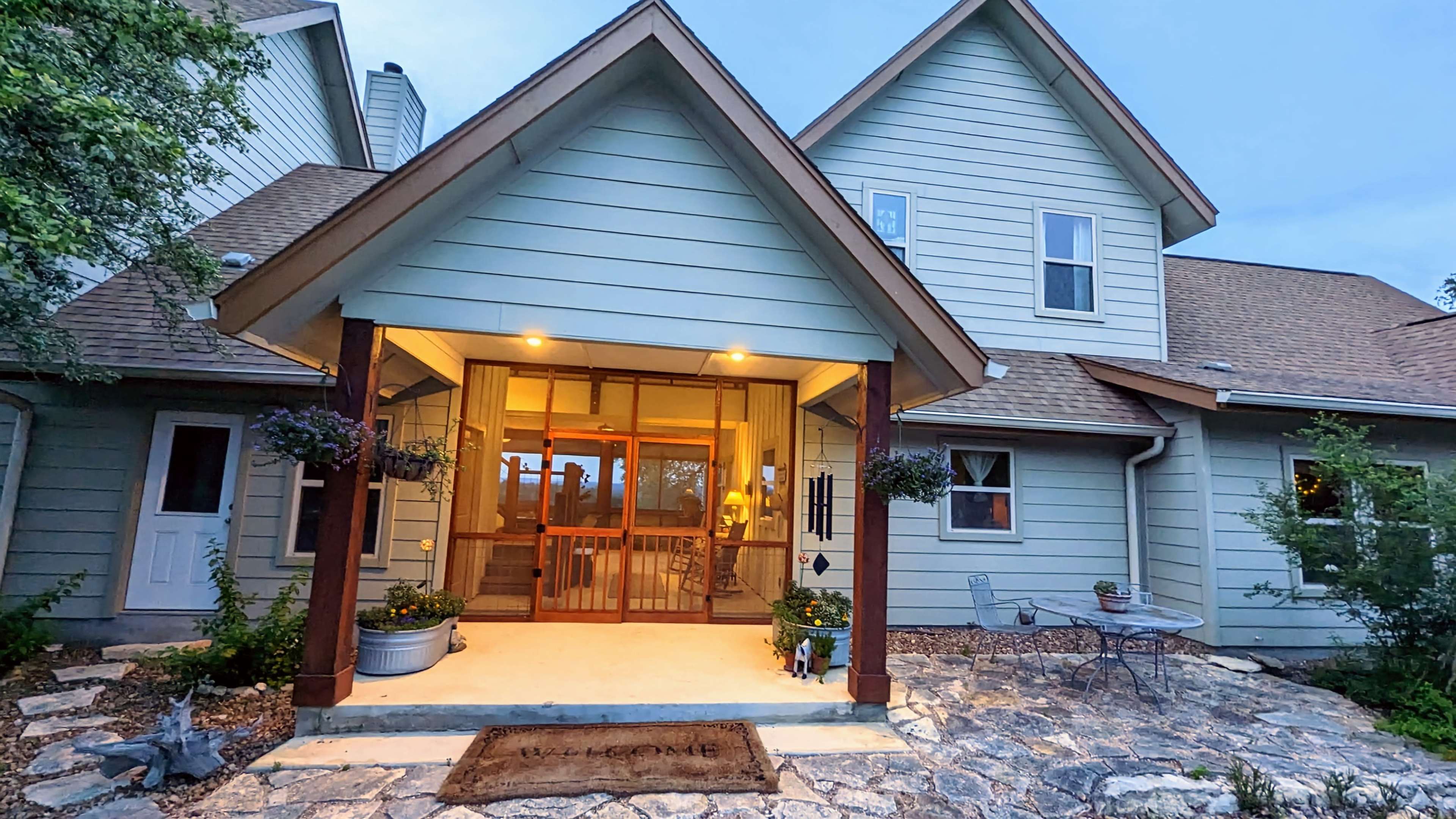 The entrance of a light blue house features a covered porch, stone walkway, and decorative potted plants.