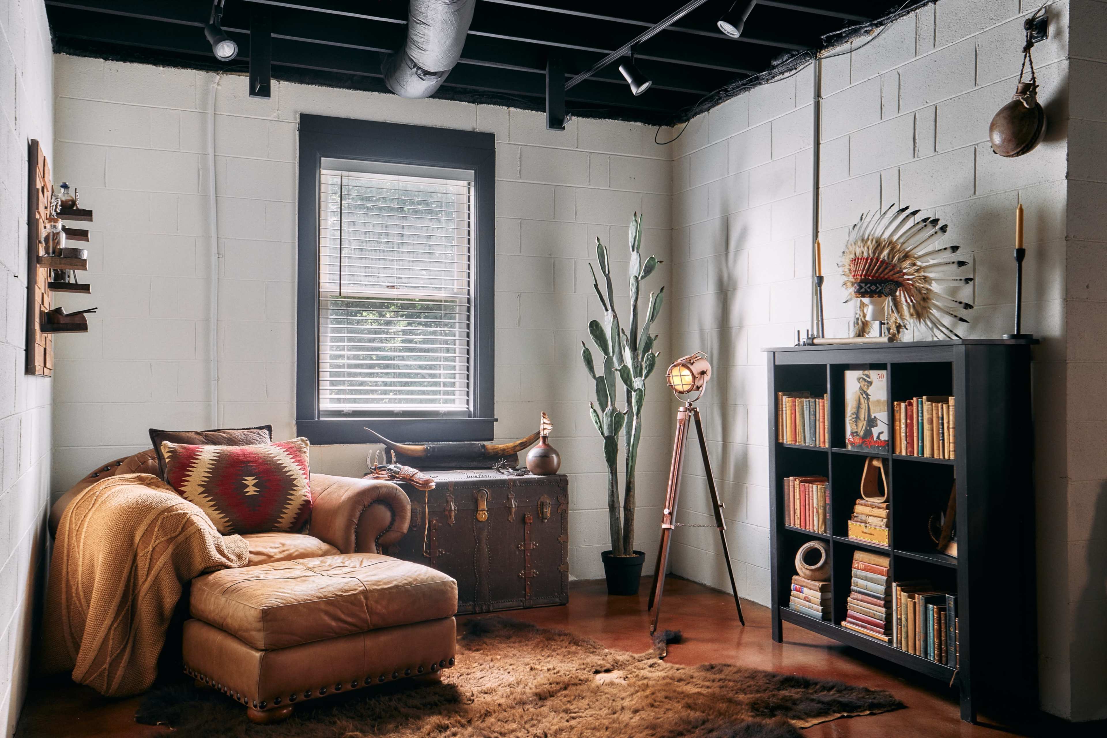 A cozy corner of a room features a leather chaise lounge, a chest, a tall cactus, and a bookshelf filled with books, accompanied by a floor lamp and decorative items.