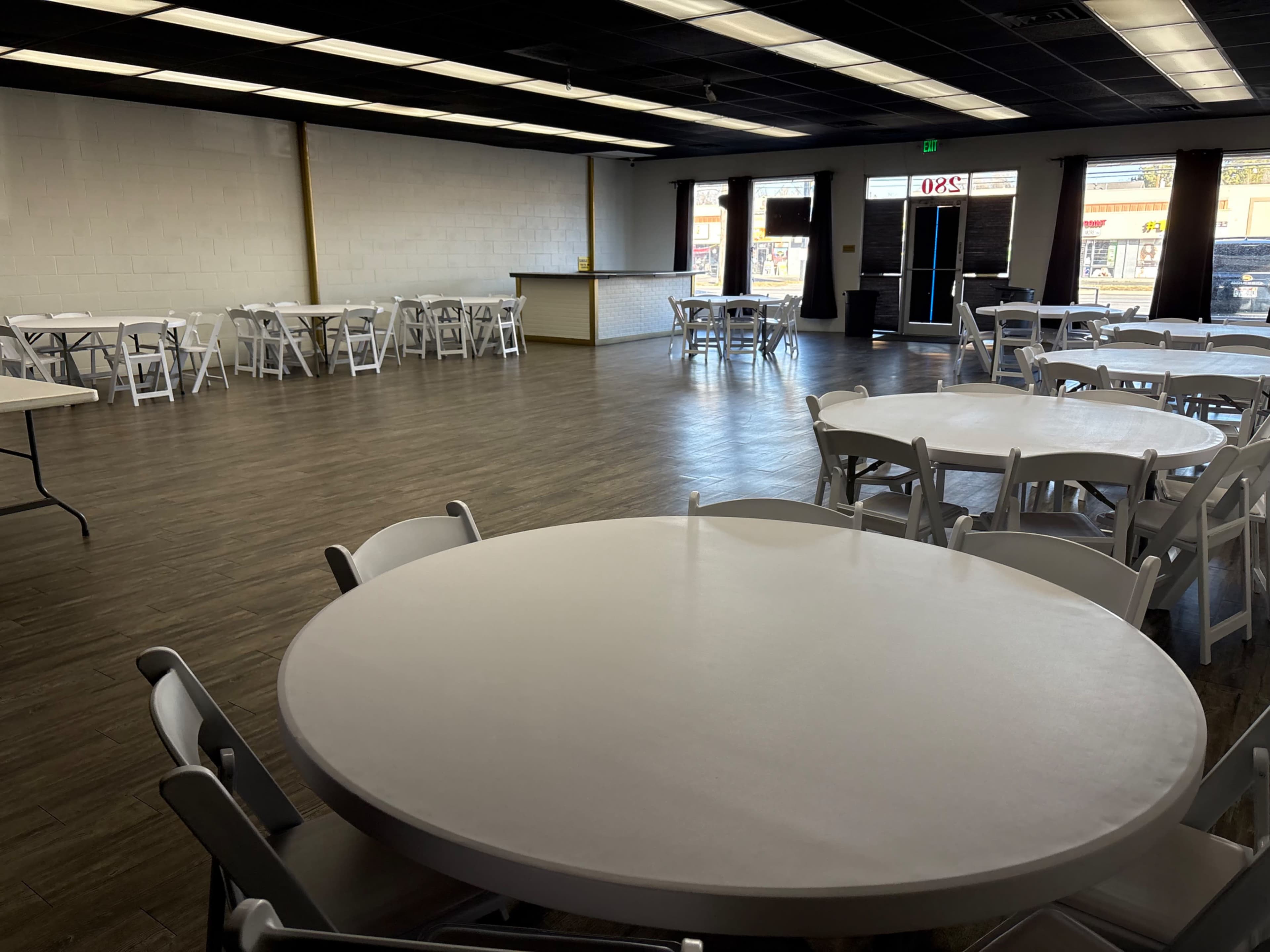 The image shows a spacious dining area with several round tables and chairs arranged neatly on a wooden floor, featuring a reception counter at the far end and large windows letting in natural light.