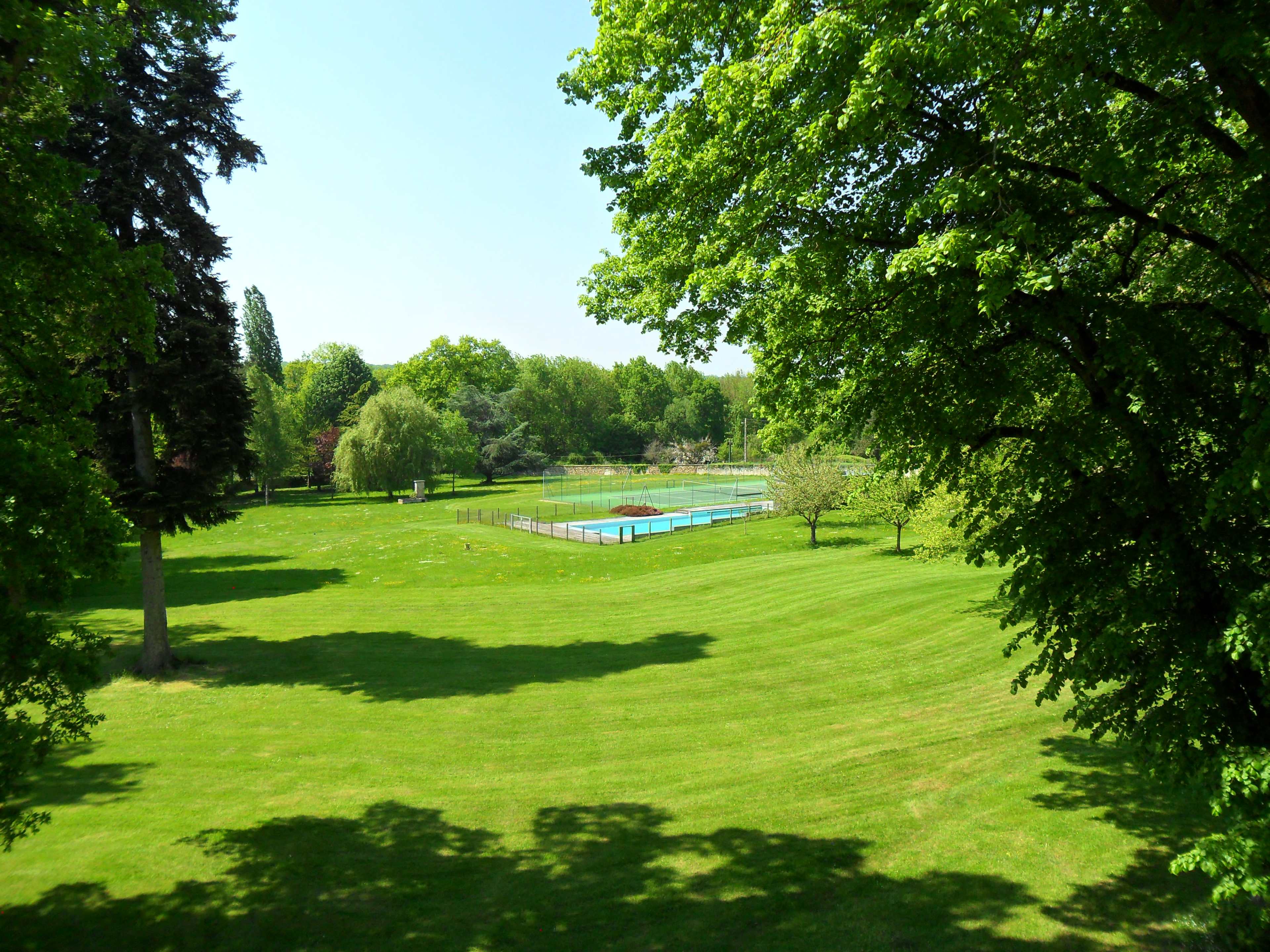 A grassy landscape features a swimming pool enclosed by a fence, surrounded by trees and greenery.