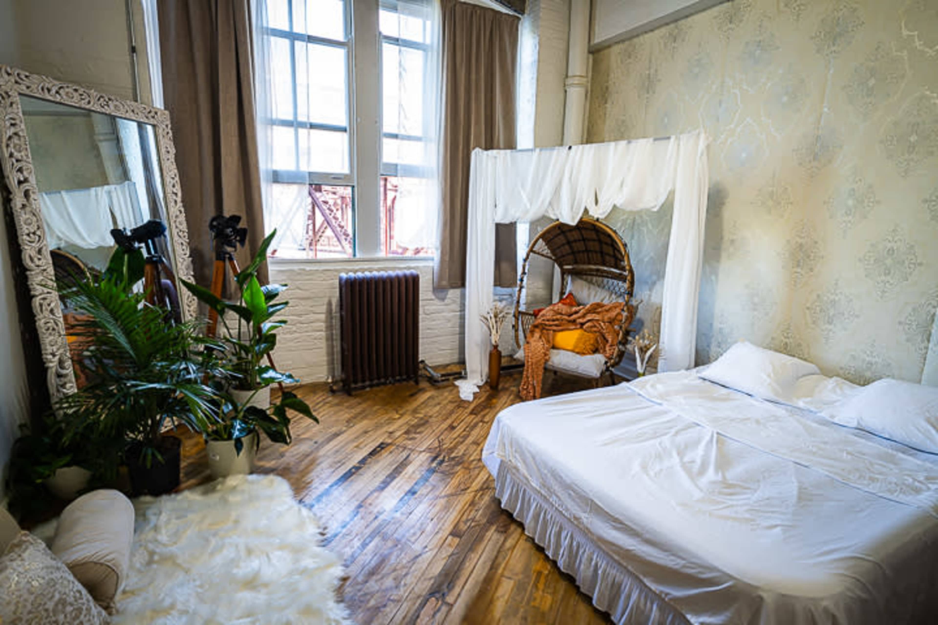 A cozy bedroom featuring a bed with white linens, a wicker seating area draped with a canopy, a large mirror, and a potted plant, all set against a backdrop of wooden floors and patterned wallpaper.