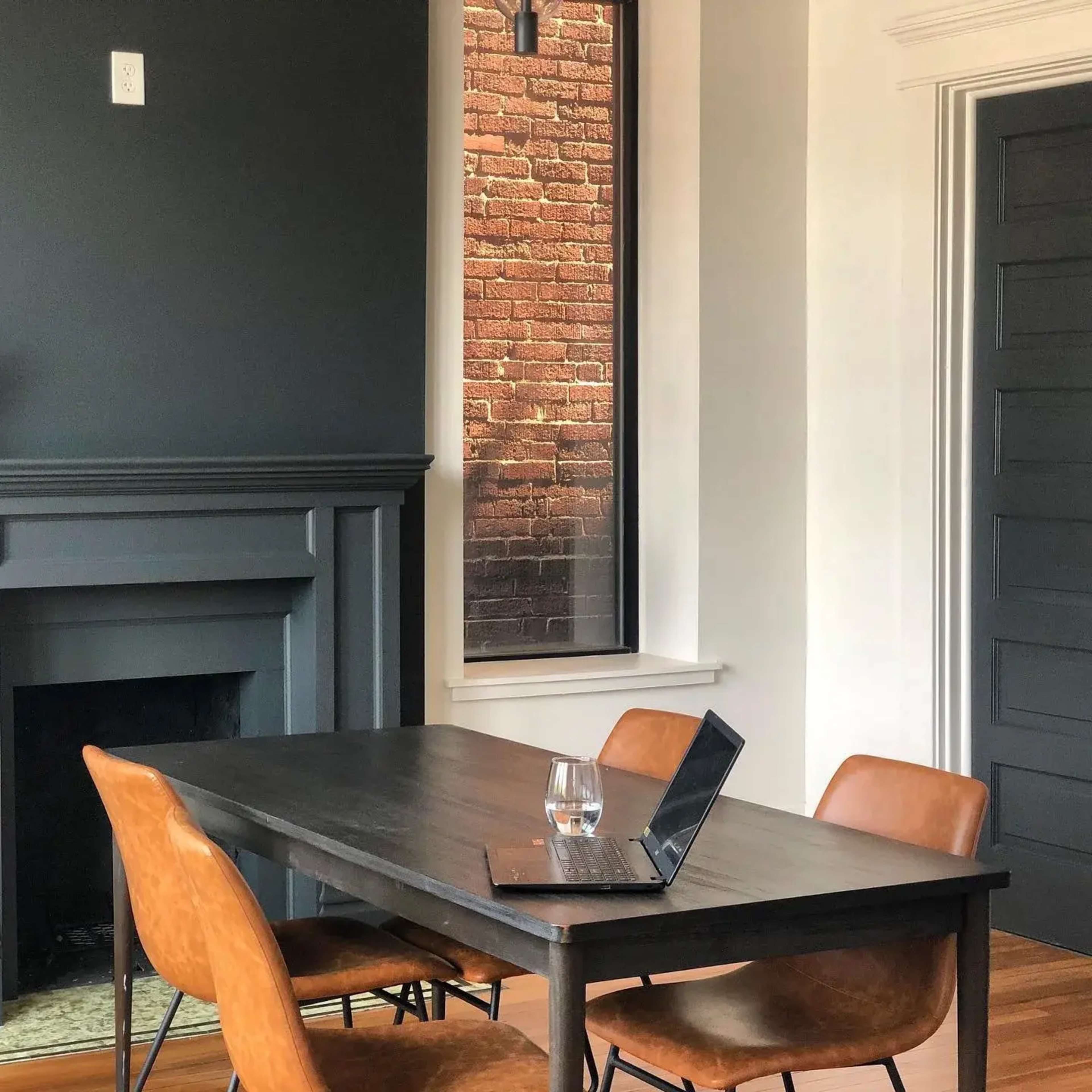 A wooden dining table with a laptop and a glass of water is positioned in a room featuring a dark wall and a large window showcasing a brick exterior.