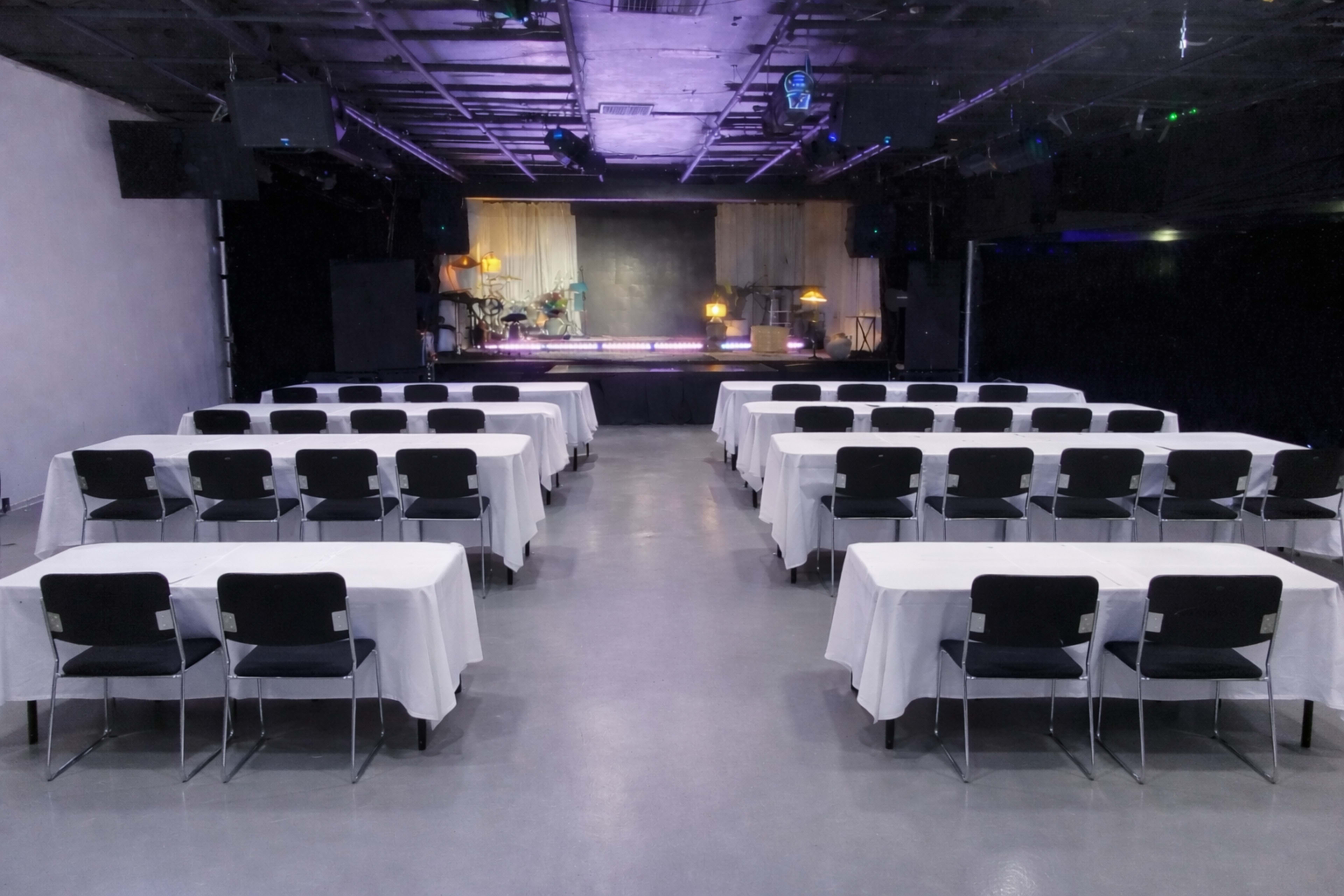 The image shows an empty event space with rows of tables covered in white tablecloths facing a stage with dim lighting.
