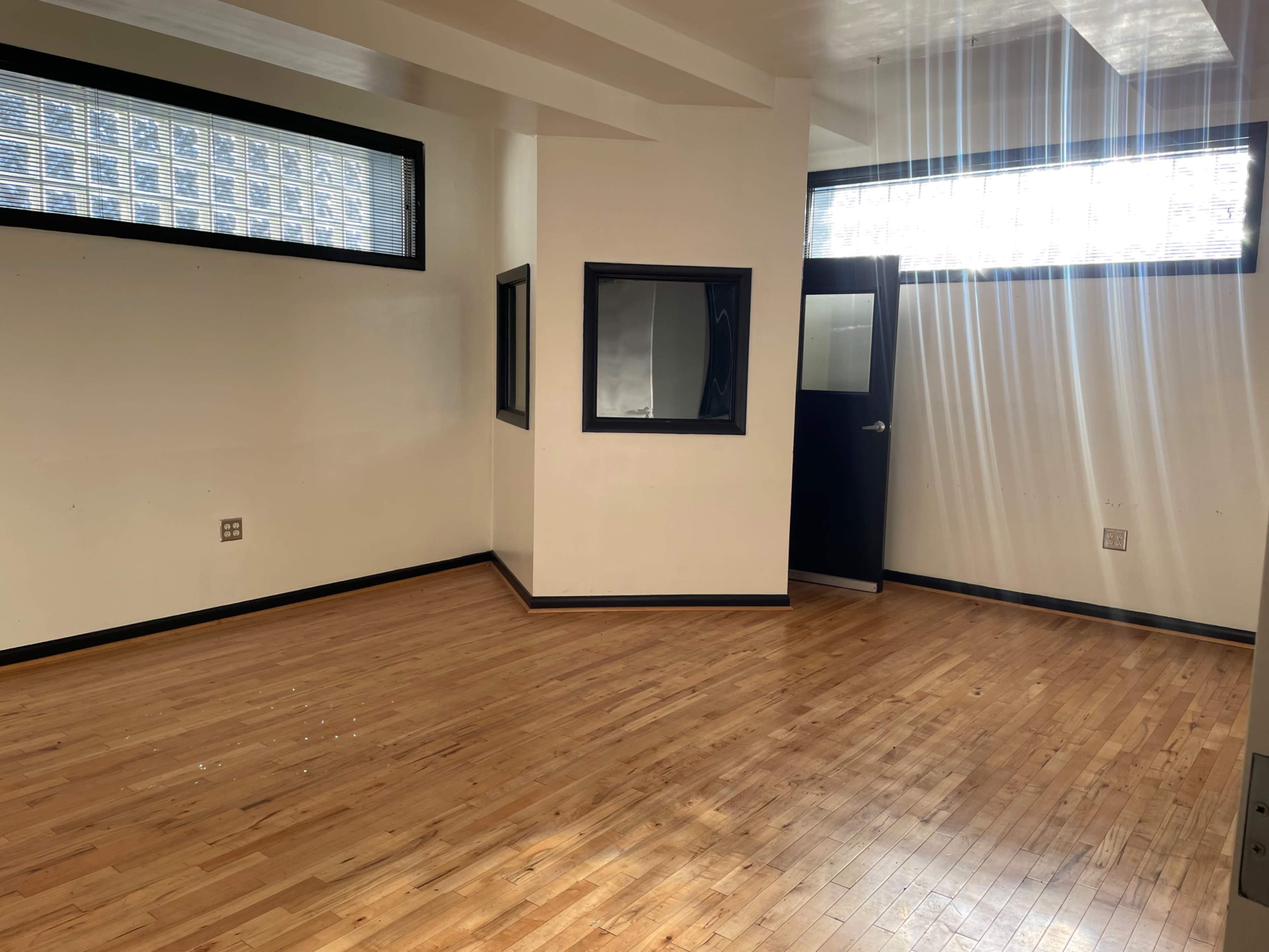 The image shows an empty room with wooden floors and large windows that allow natural light to enter.