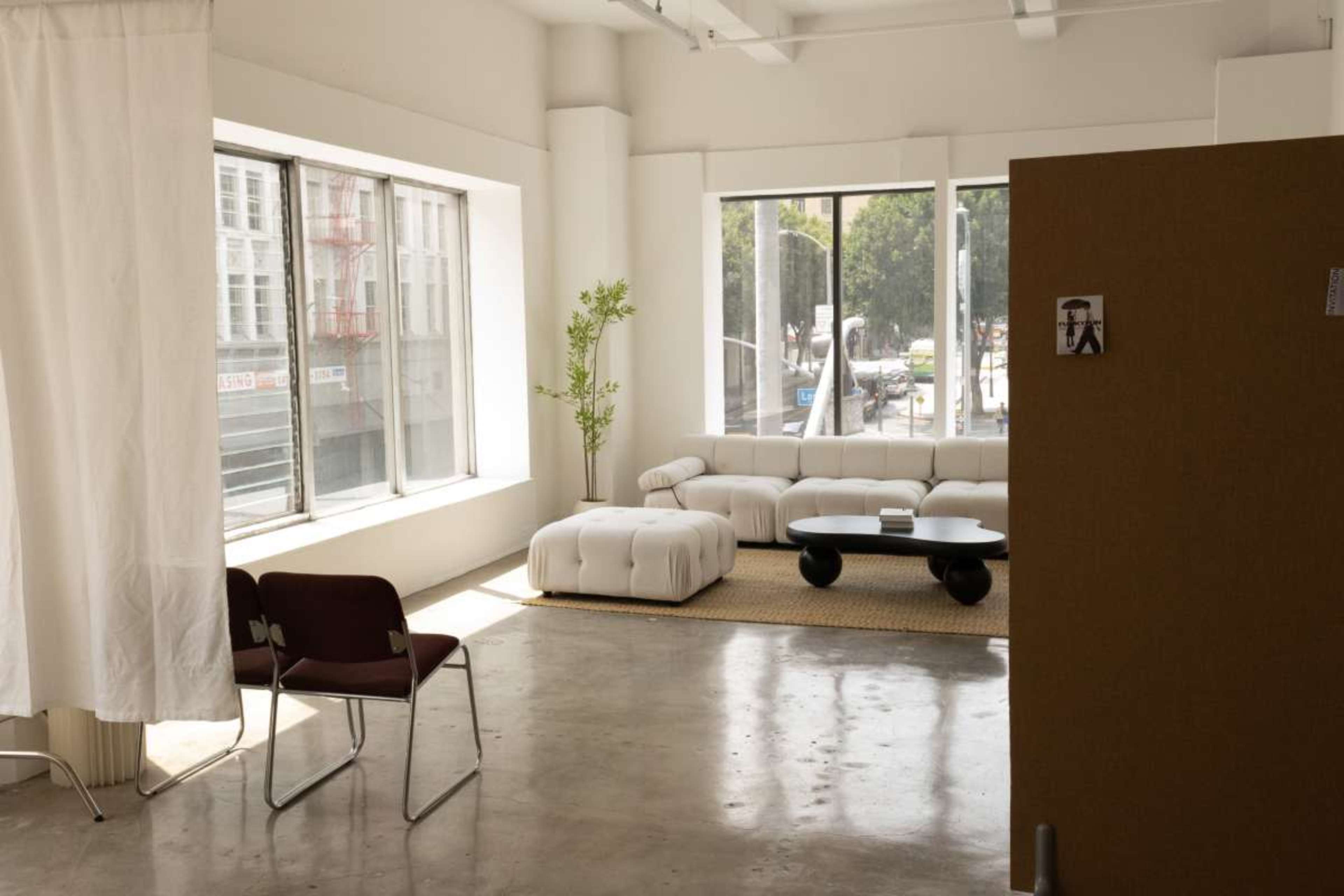 The image shows a bright, contemporary space with a white sectional couch, a round black coffee table, and large windows allowing natural light to enter.