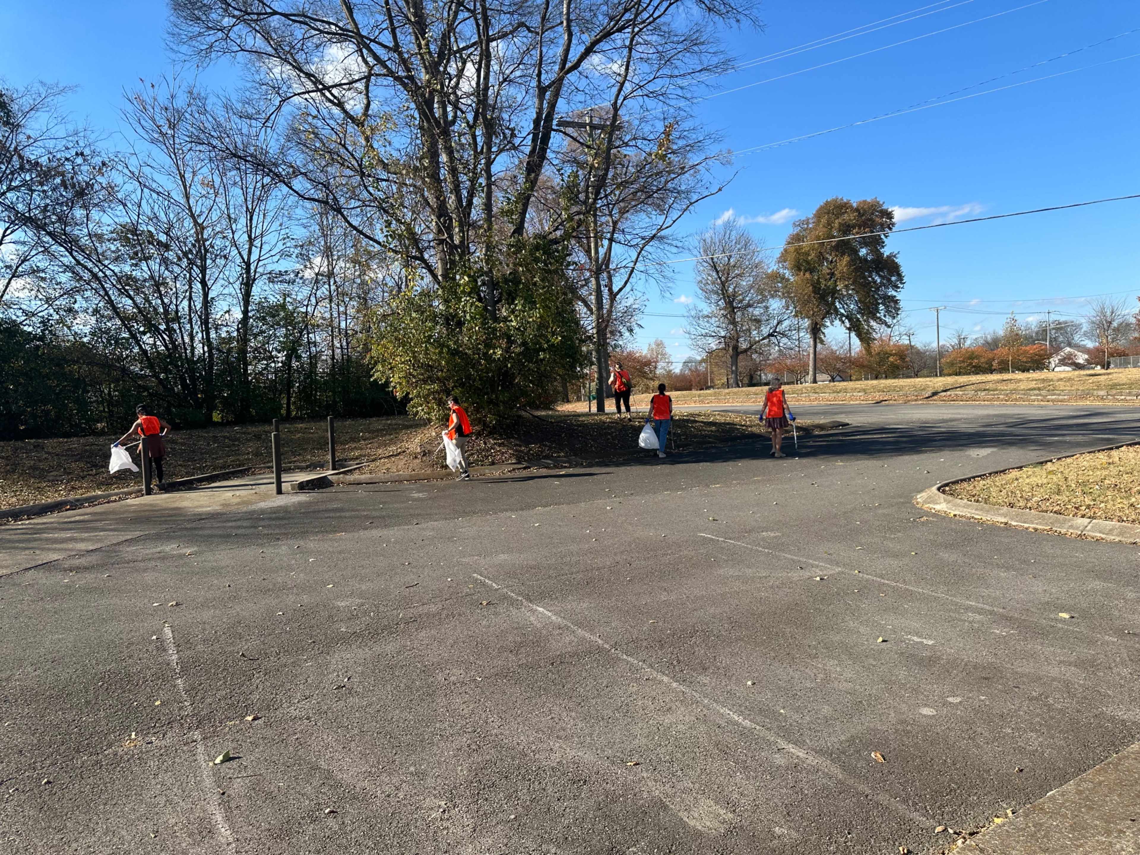 A group of people in orange shirts collects trash along a roadside near a park.