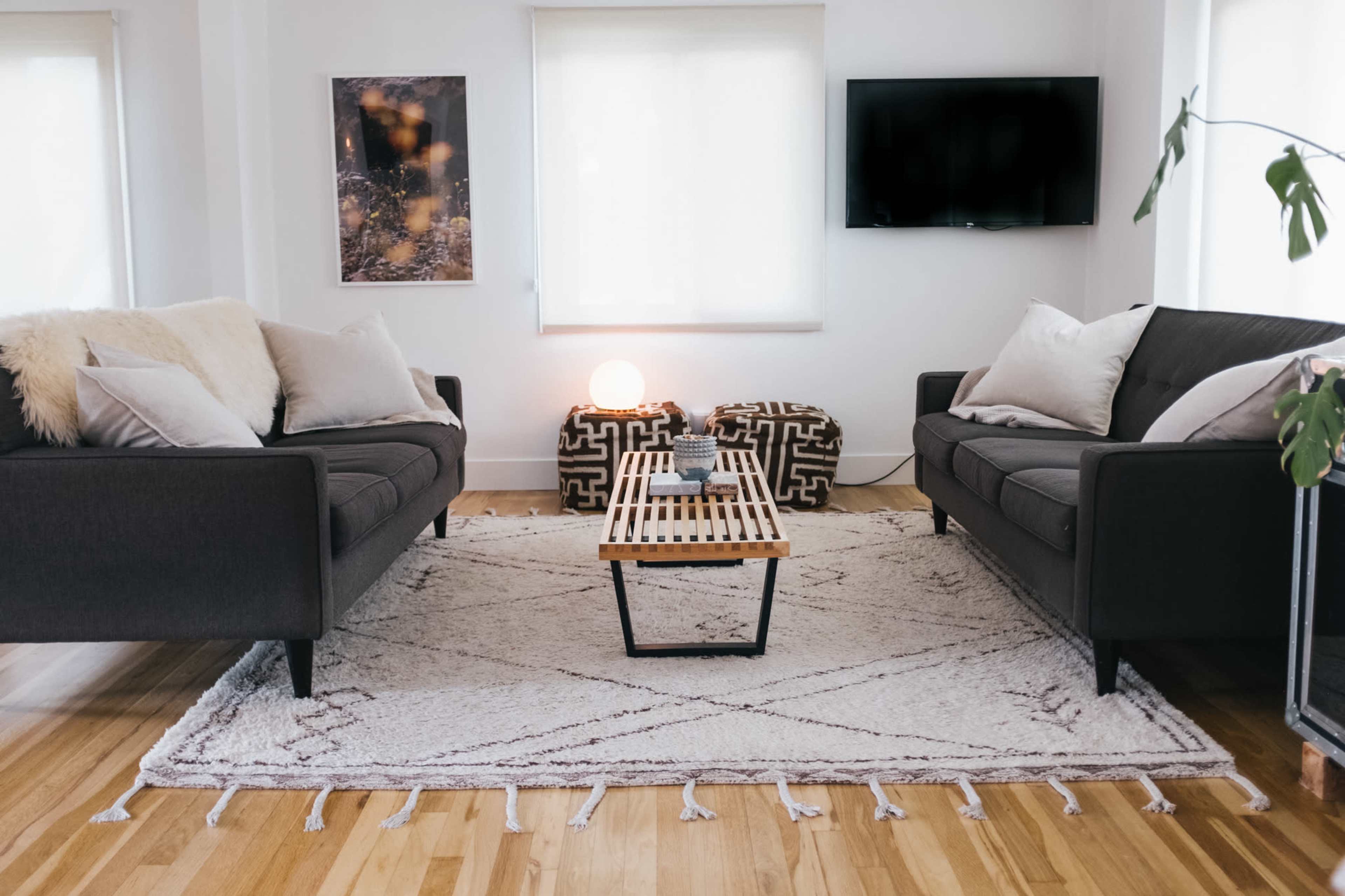 A modern living room with two dark sofas facing a coffee table on a patterned rug, a television mounted on the wall, and large windows letting in natural light.