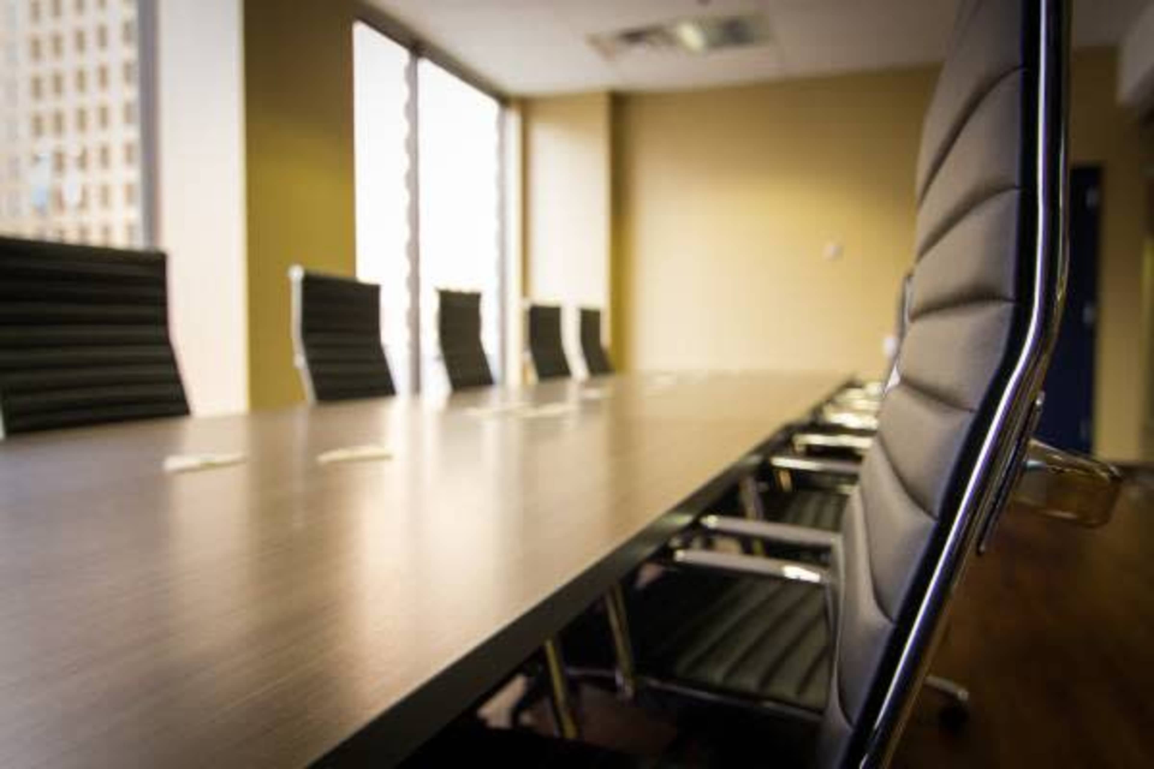 A dimly lit conference room features a long, wooden table surrounded by black leather chairs, with large windows letting in natural light.