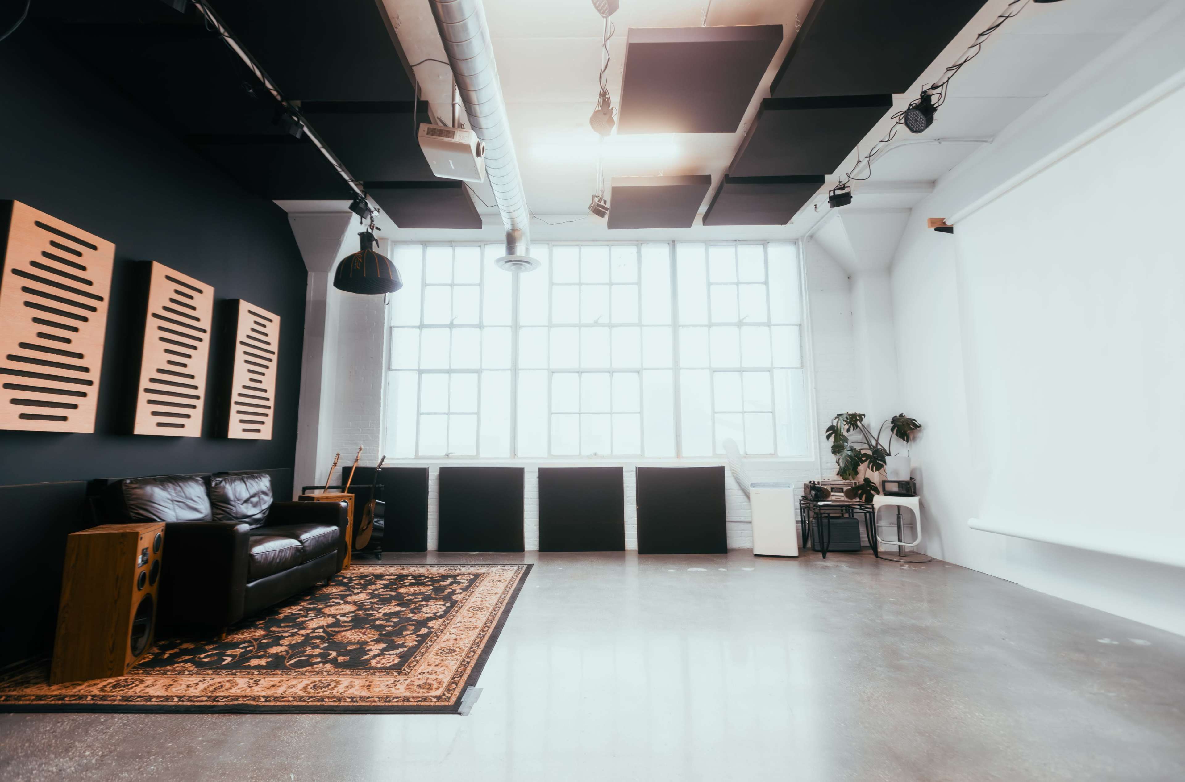 The image shows a spacious, modern studio with a leather couch, large windows, and a decorative rug on polished concrete flooring.