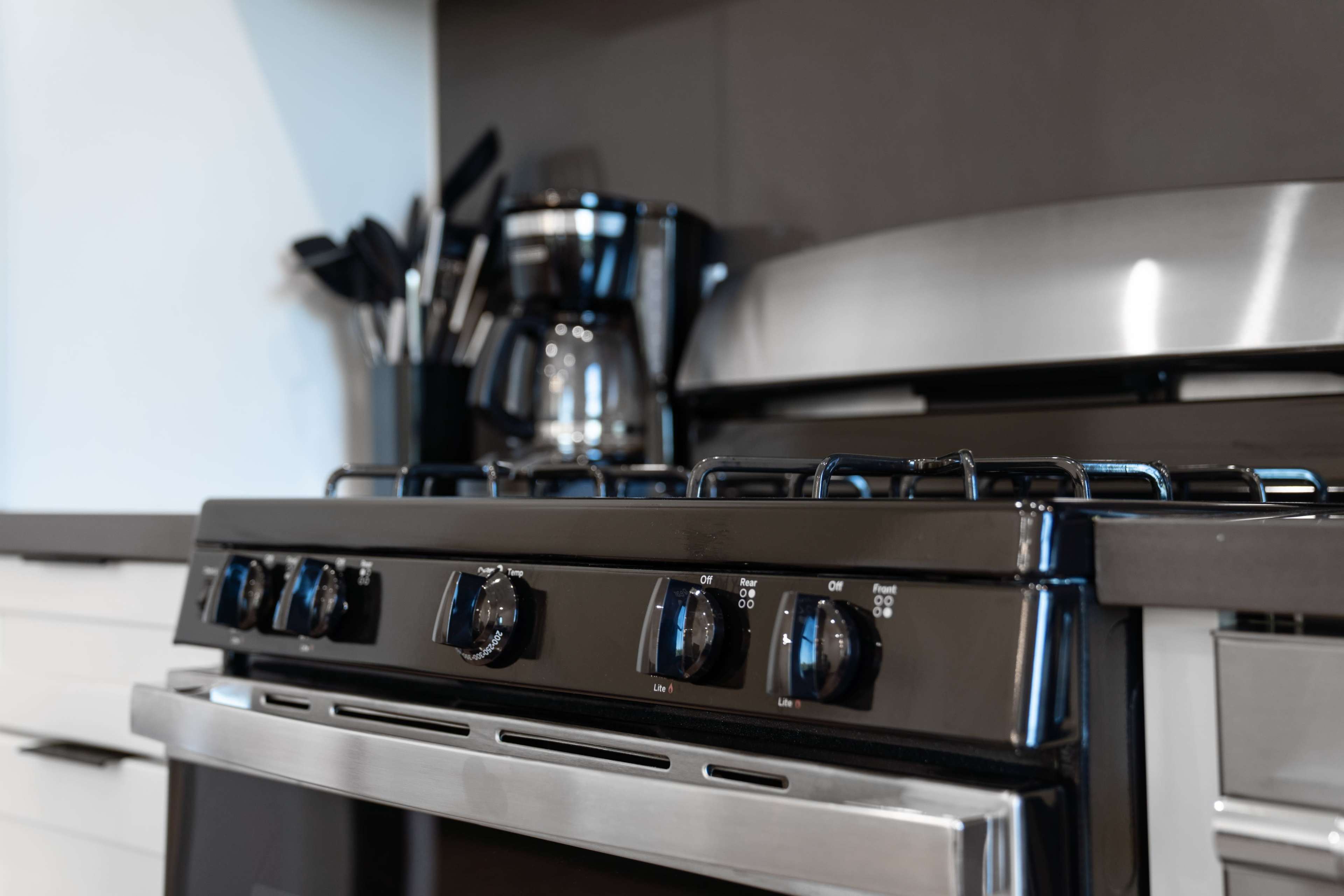 A black gas stove is positioned next to a silver countertop and kitchen utensils, with a coffee maker in the background.