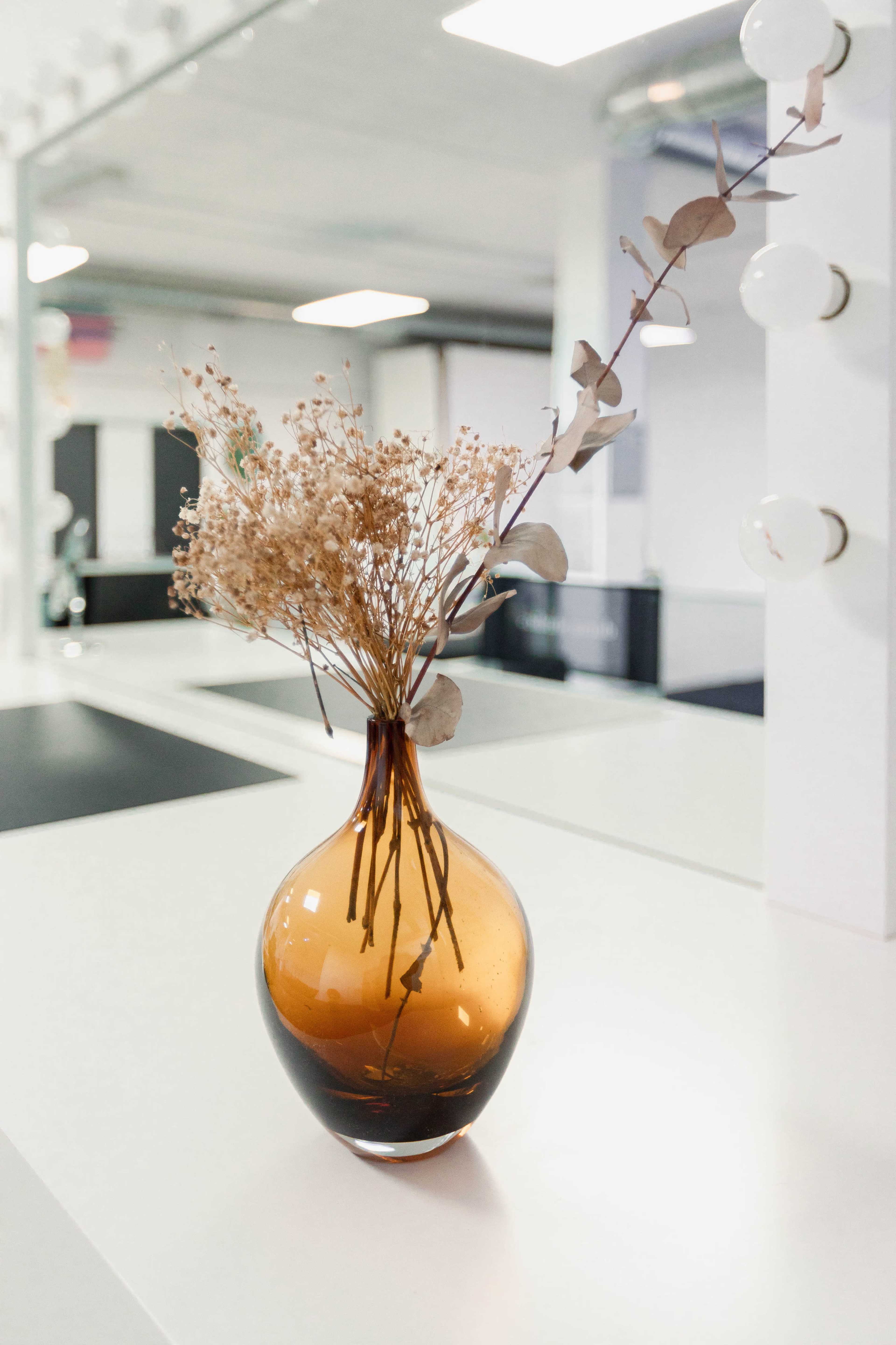 A brown glass vase holds dried flowers and branches on a white table in a bright room.
