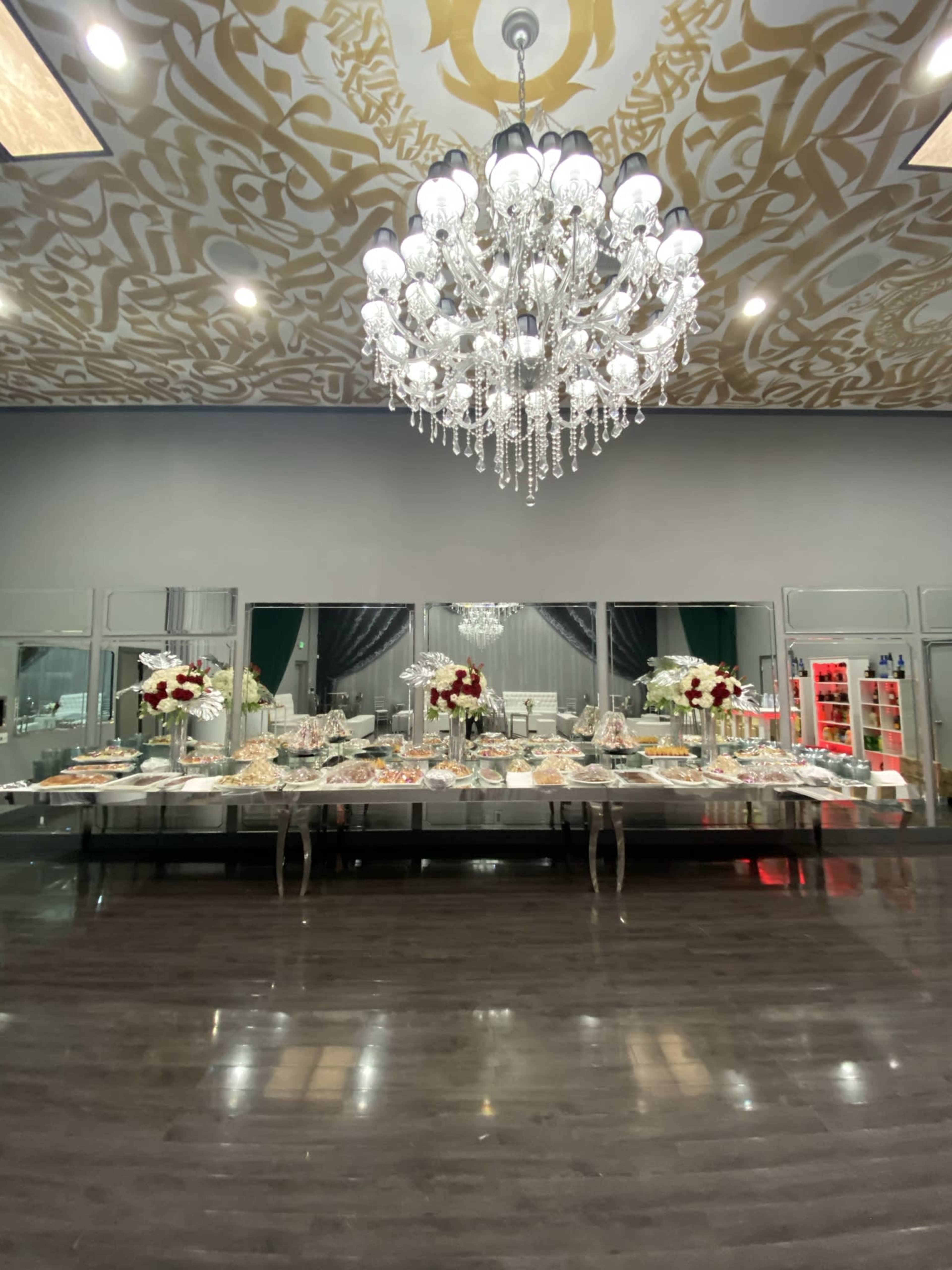 An elegantly decorated banquet hall featuring a large buffet table adorned with floral arrangements and a crystal chandelier overhead.