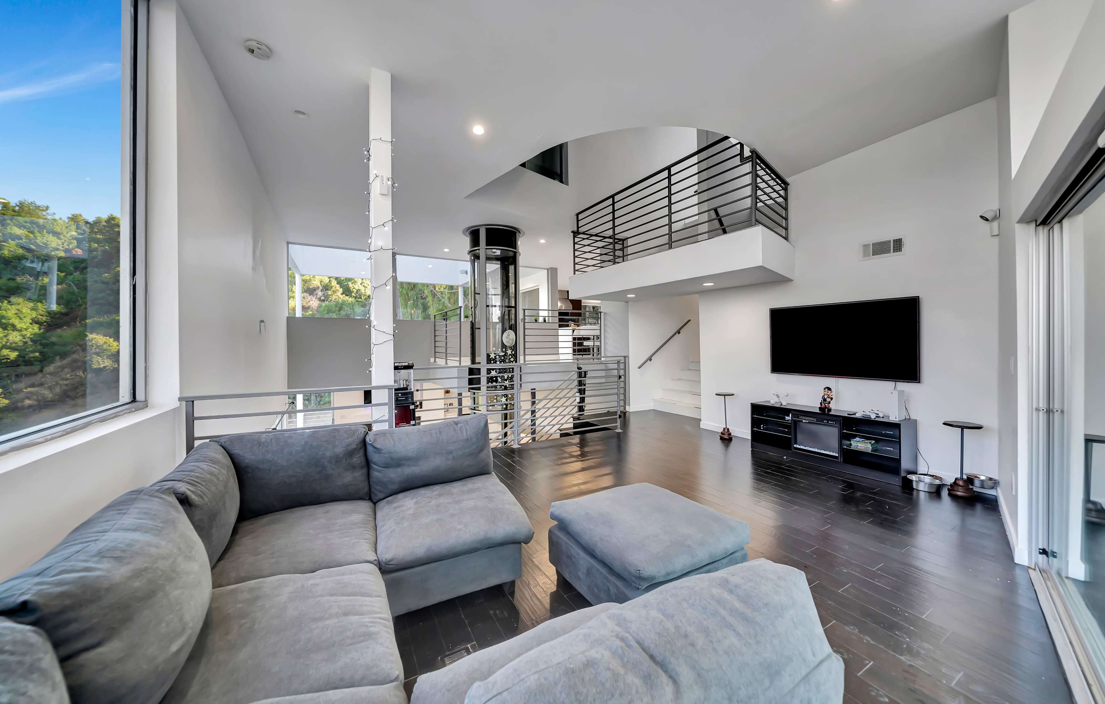 A modern living room with a gray sectional sofa, large windows, and a staircase leading to an upper level.
