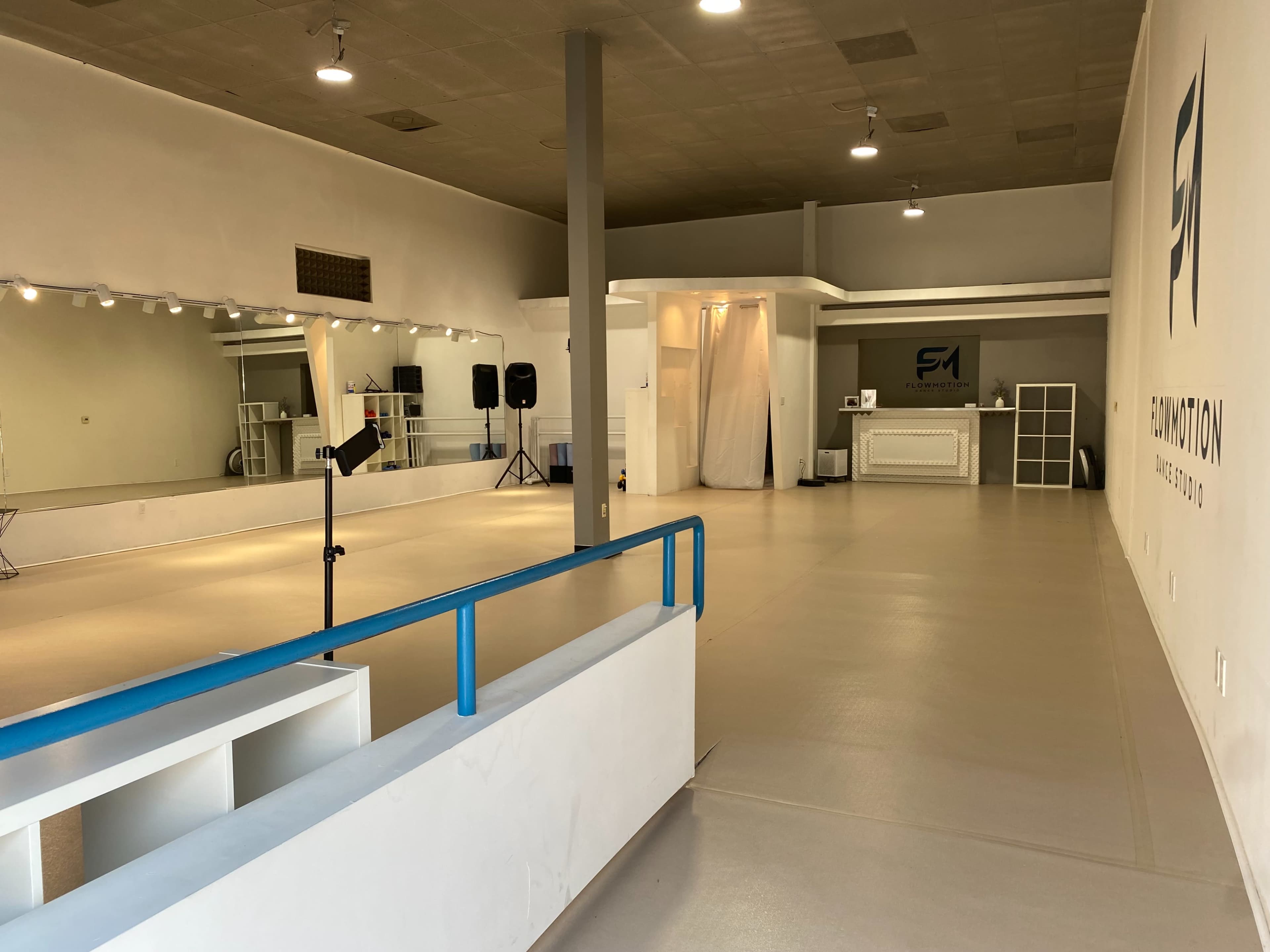 A spacious dance studio with a mirrored wall, a music stand, and a reception area featuring white decor and a logo on the wall.