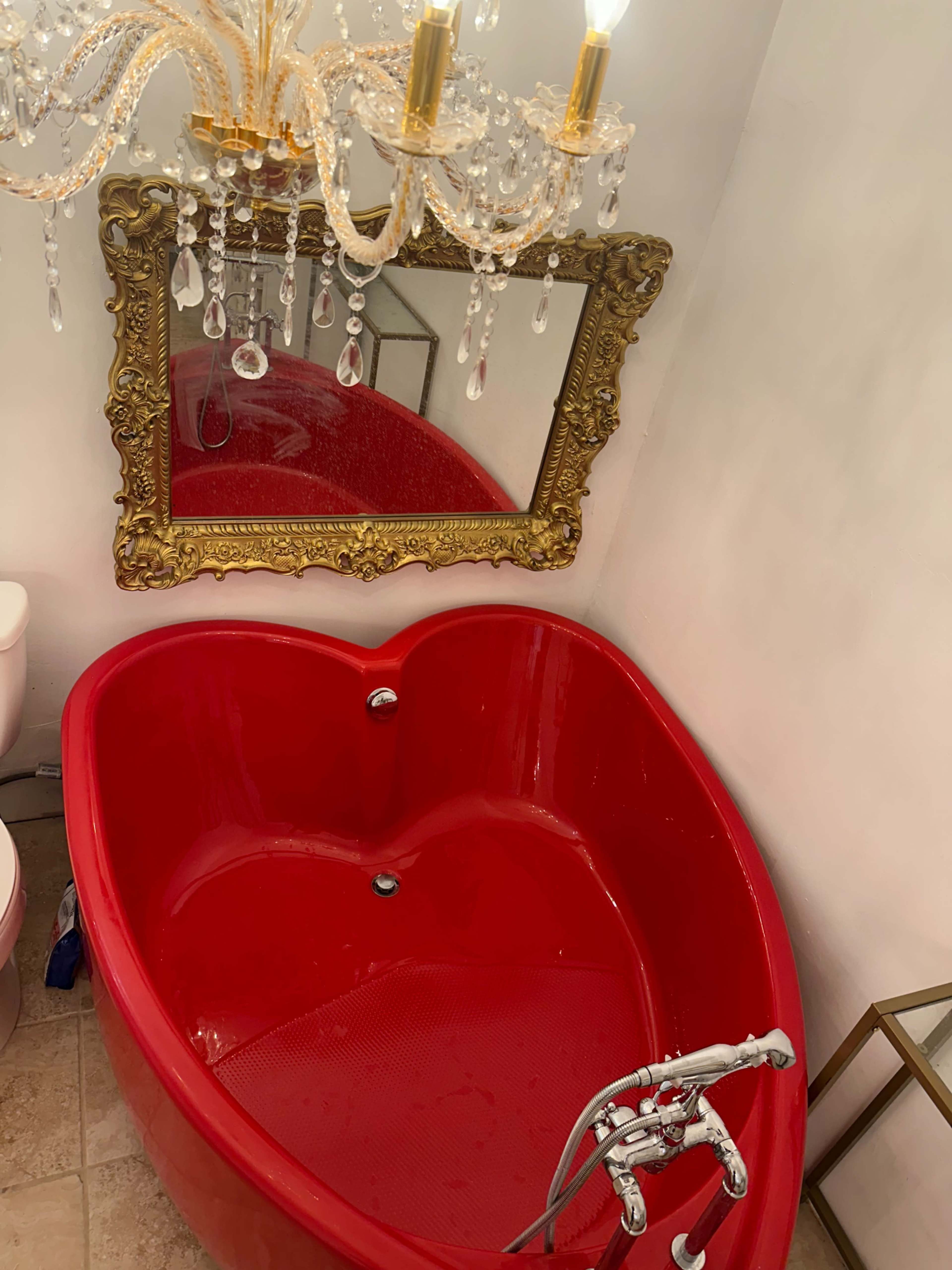 A red heart-shaped bathtub is positioned beneath a ornate gold-framed mirror and a crystal chandelier in a bathroom setting.