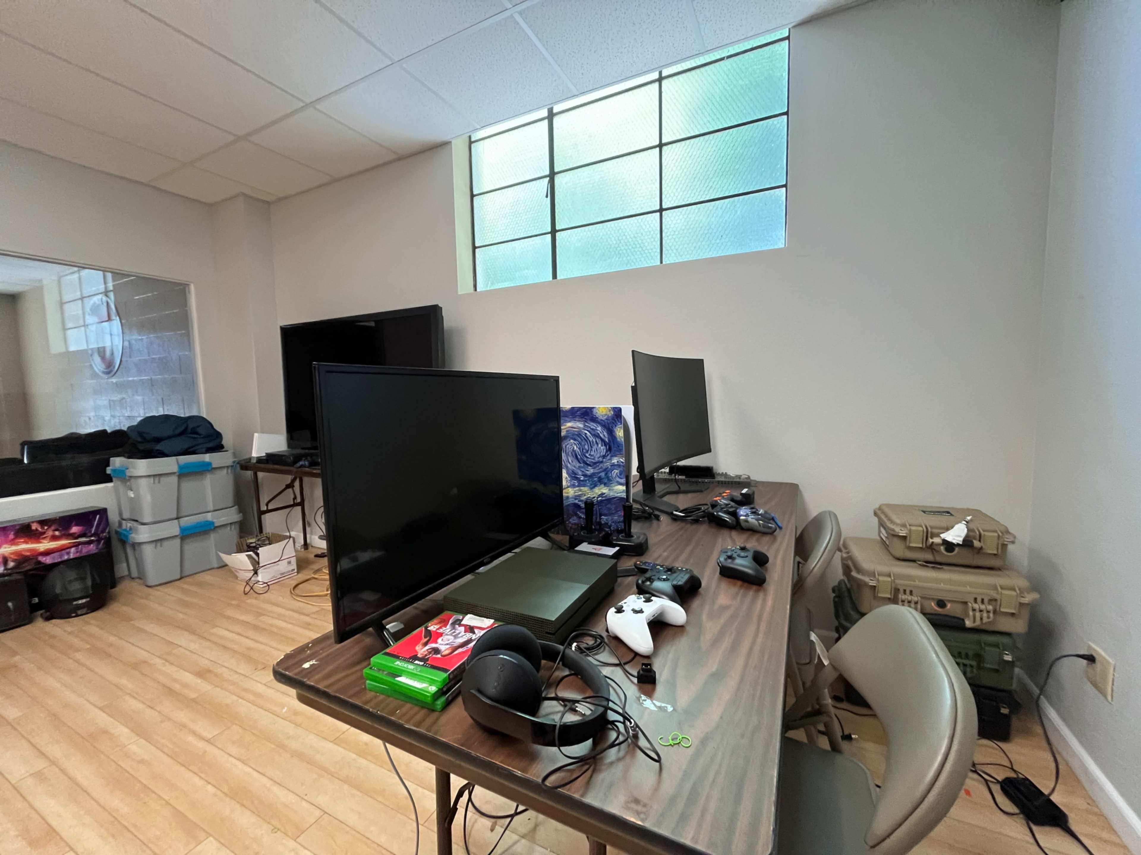 The image shows a room with a table holding two gaming consoles, a large TV, and various gaming accessories, with a window in the background.
