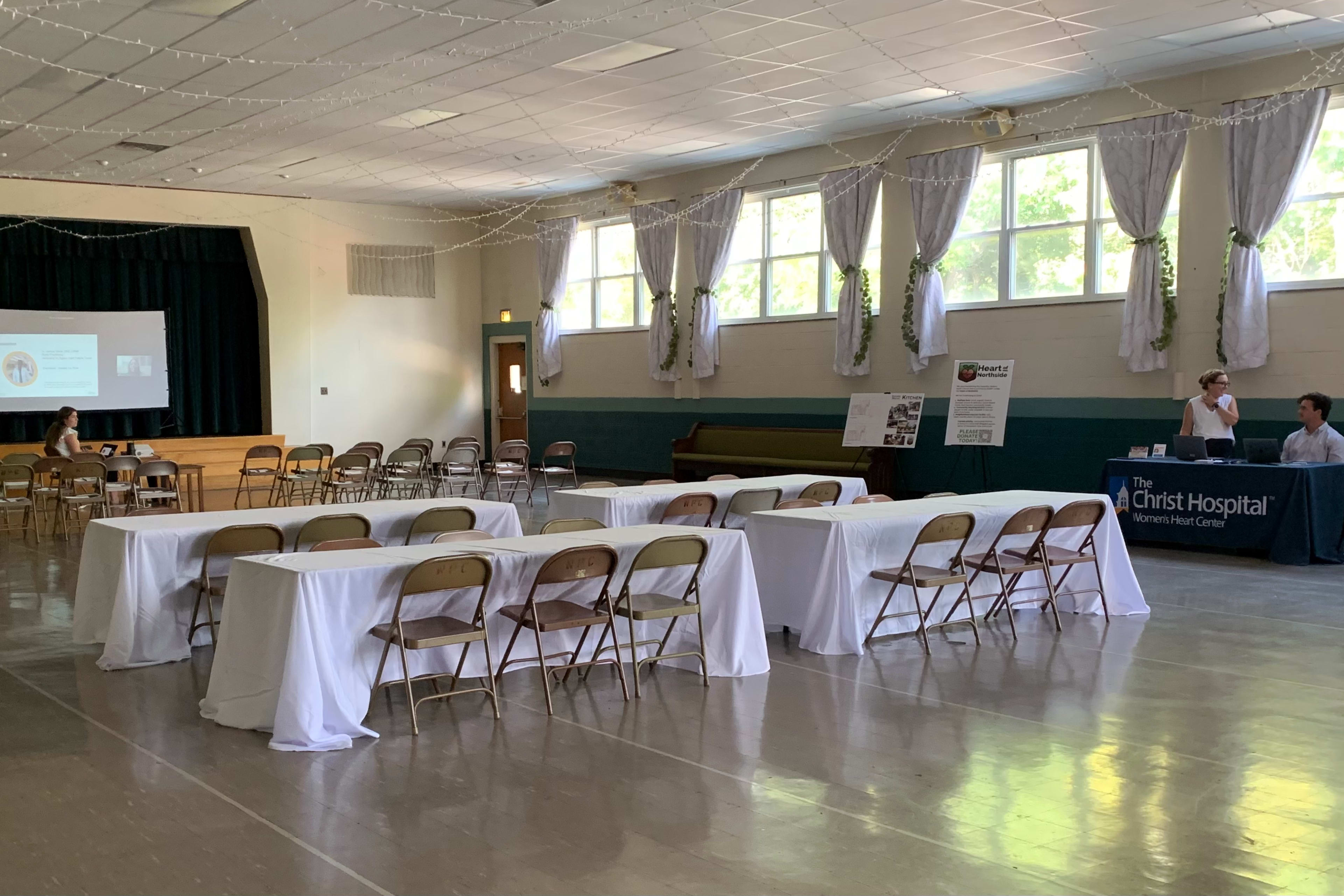 A spacious indoor venue features rows of chairs and tables, with a projector screen in the background and a booth set up in a corner.