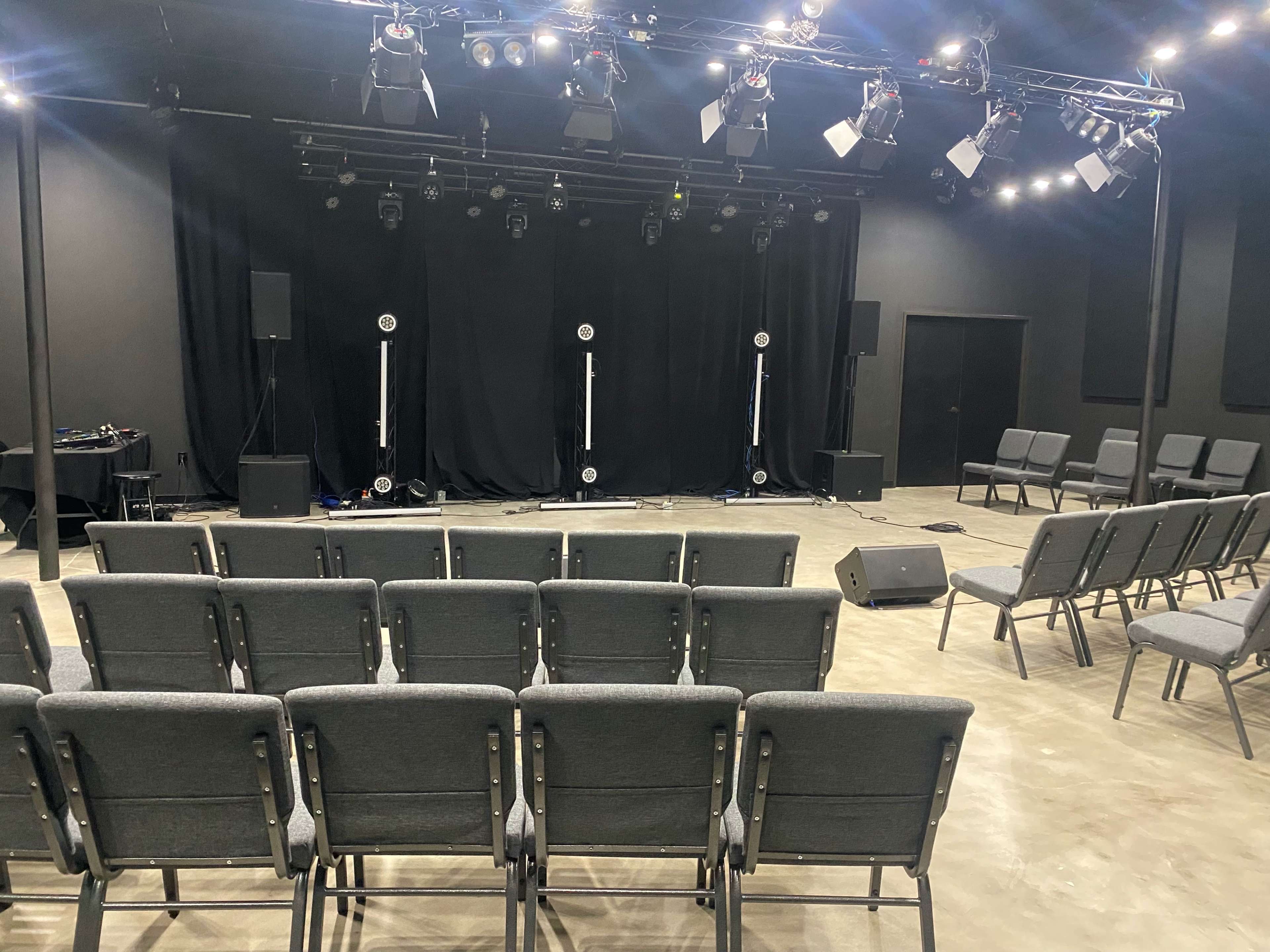 A stage with black curtains and lighting equipment is set up in an empty performance venue, facing rows of grey chairs for the audience.