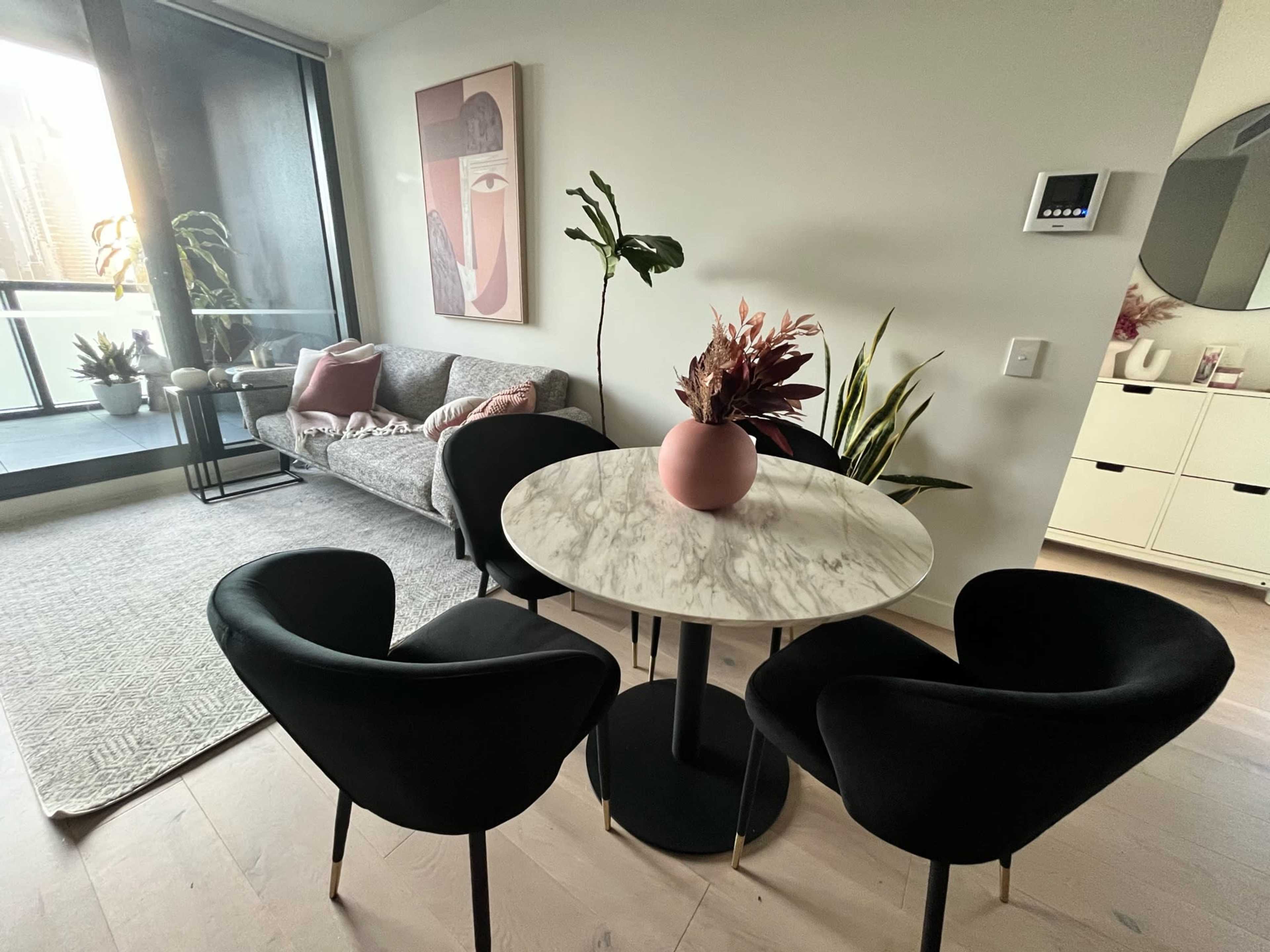 A modern living space featuring a marble dining table surrounded by four black chairs, with a gray sofa and decorative plants in the background.