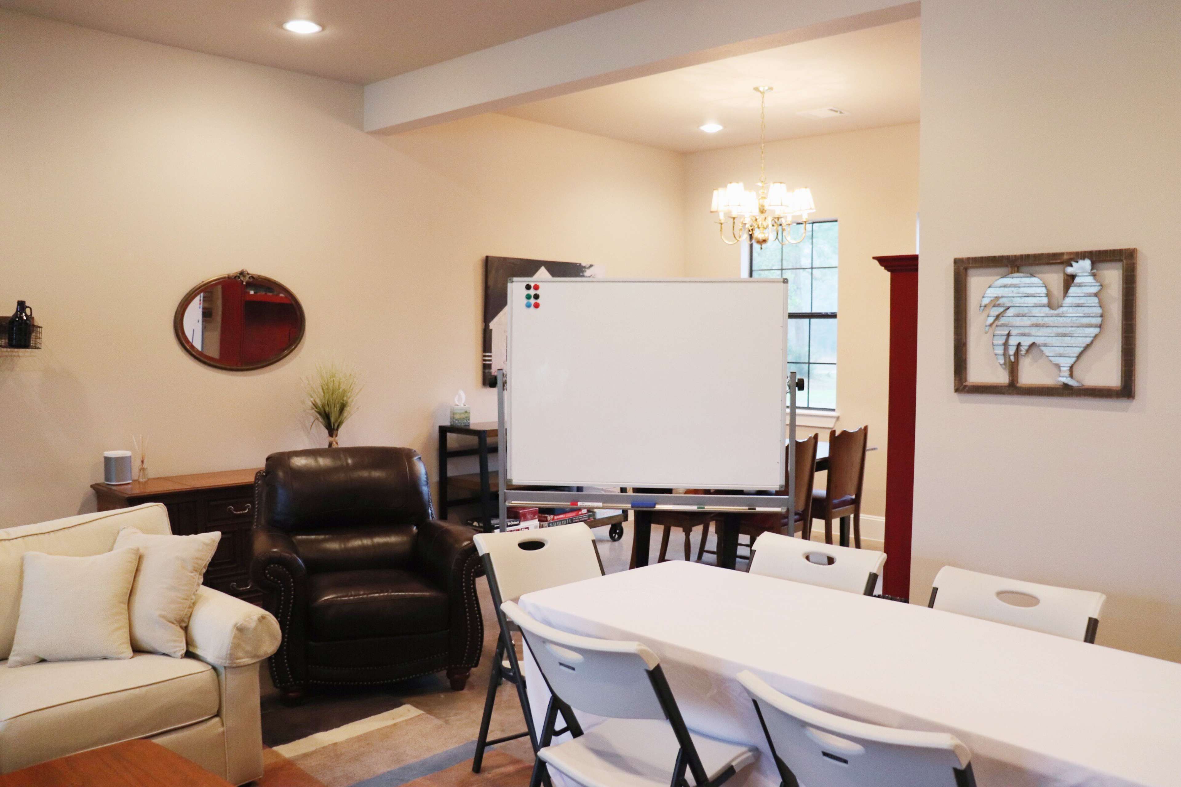 The room features a whiteboard in the center, a black leather chair, a dining area with a chandelier, and a long table with white chairs.
