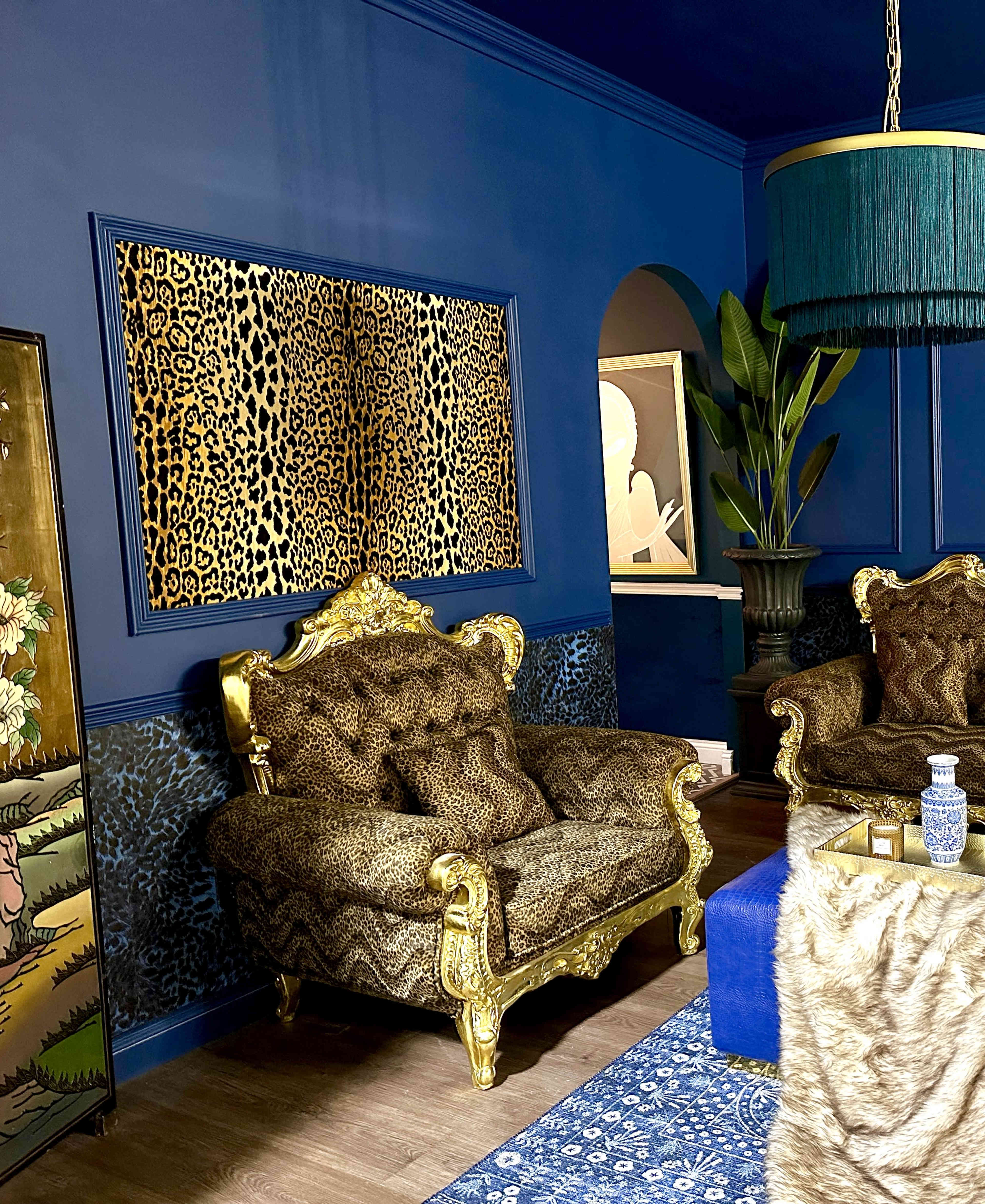 A room with dark blue walls, leopard print upholstery on two ornate armchairs, a large decorative plant, and abstract art on the wall.