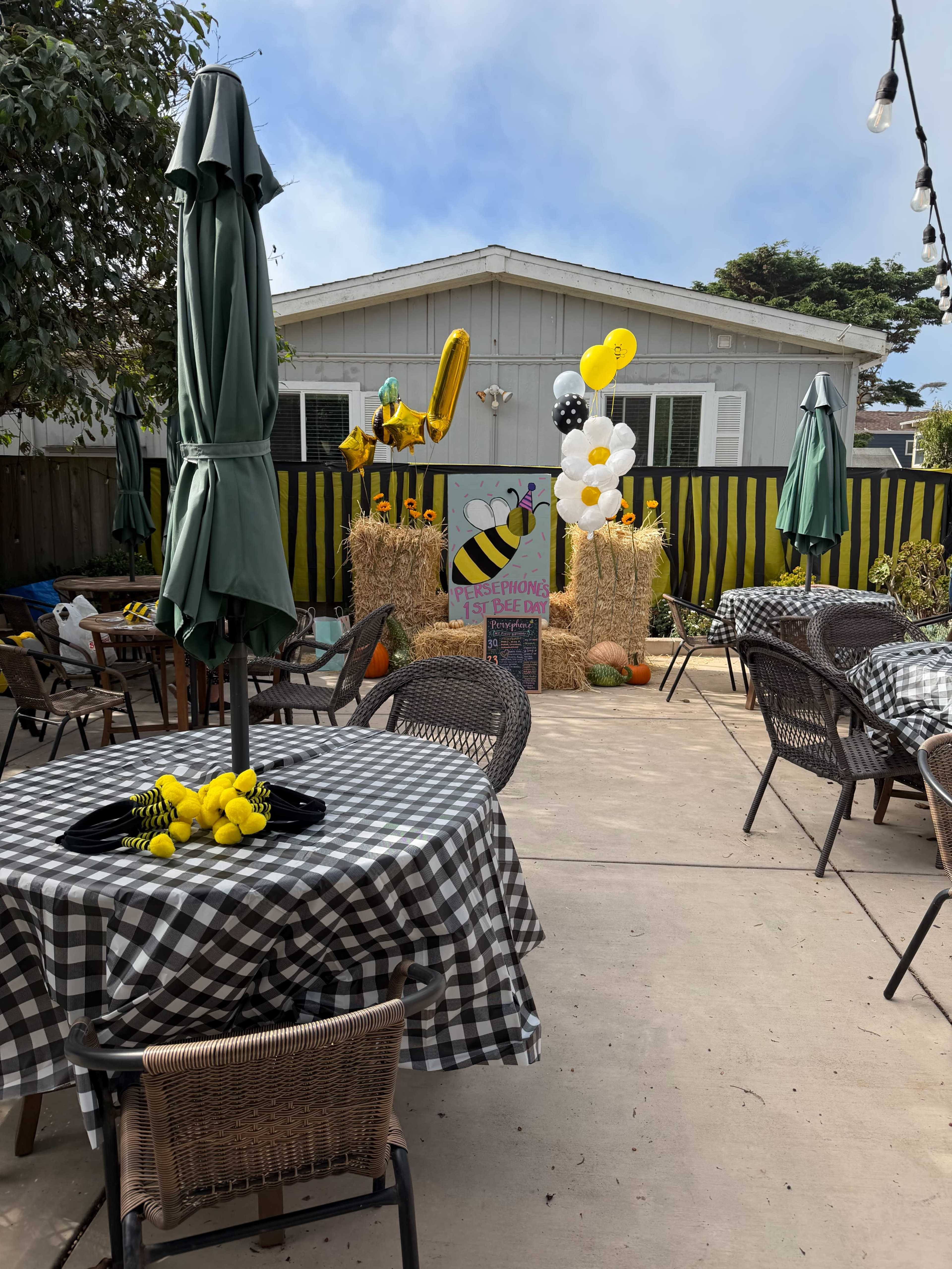 A backyard party setup features checkered tables, green umbrellas, hay bales, and colorful balloons against a house with decorative yellow accents.