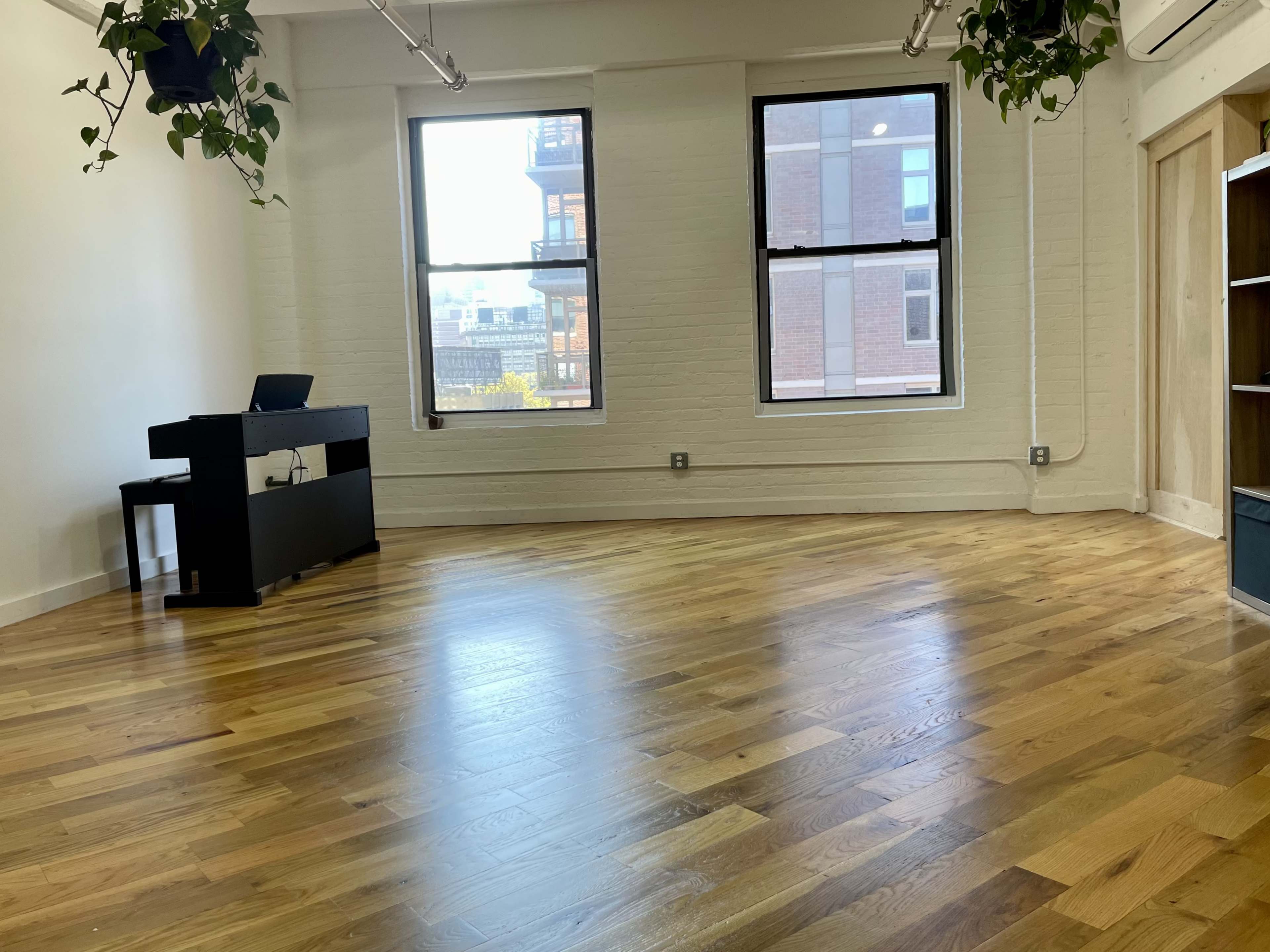A room with wooden floors features two large windows, a small black desk, and a bookshelf against one wall.