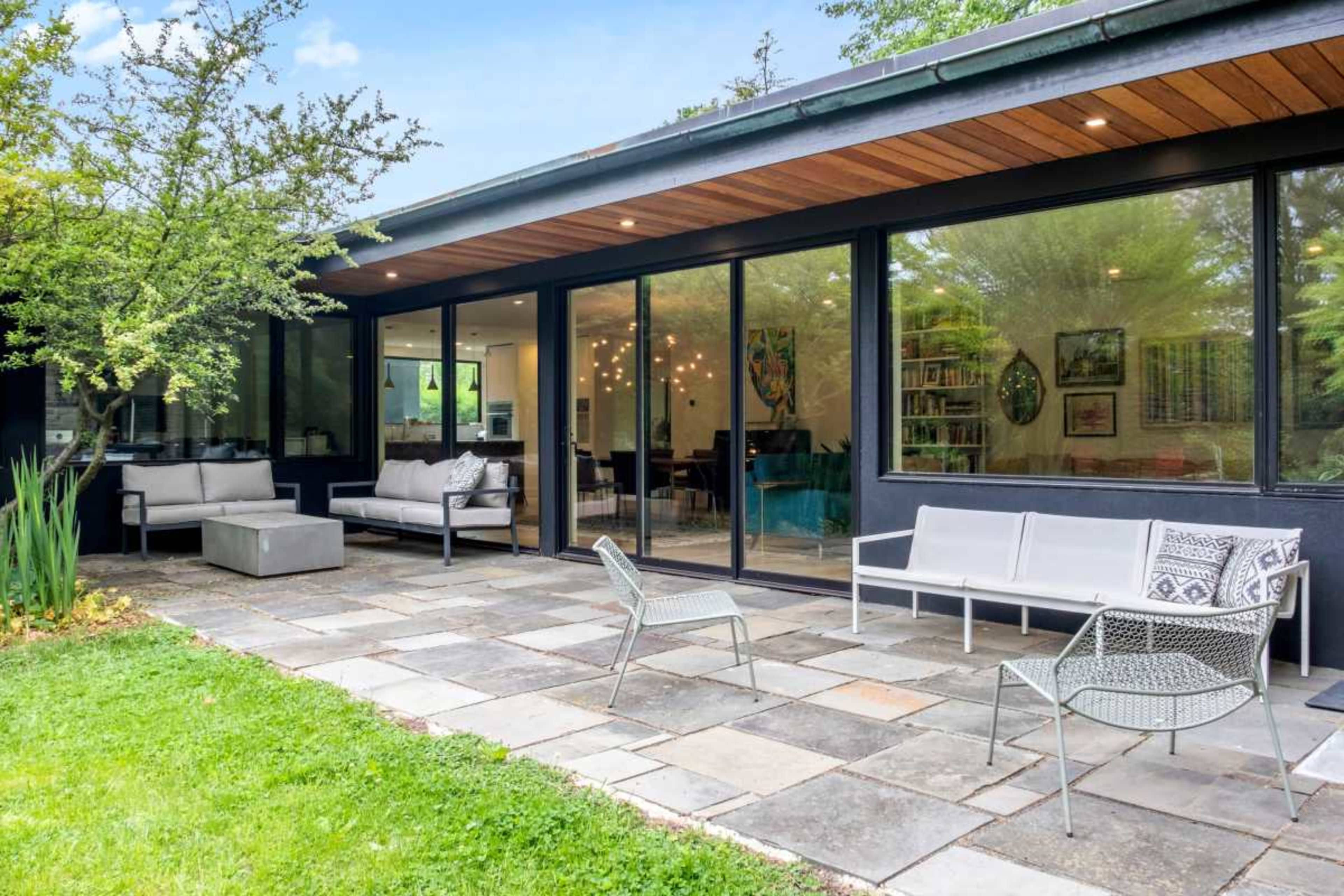 The image shows a modern patio area with stone flooring, outdoor seating, and large glass windows that open to a living space.