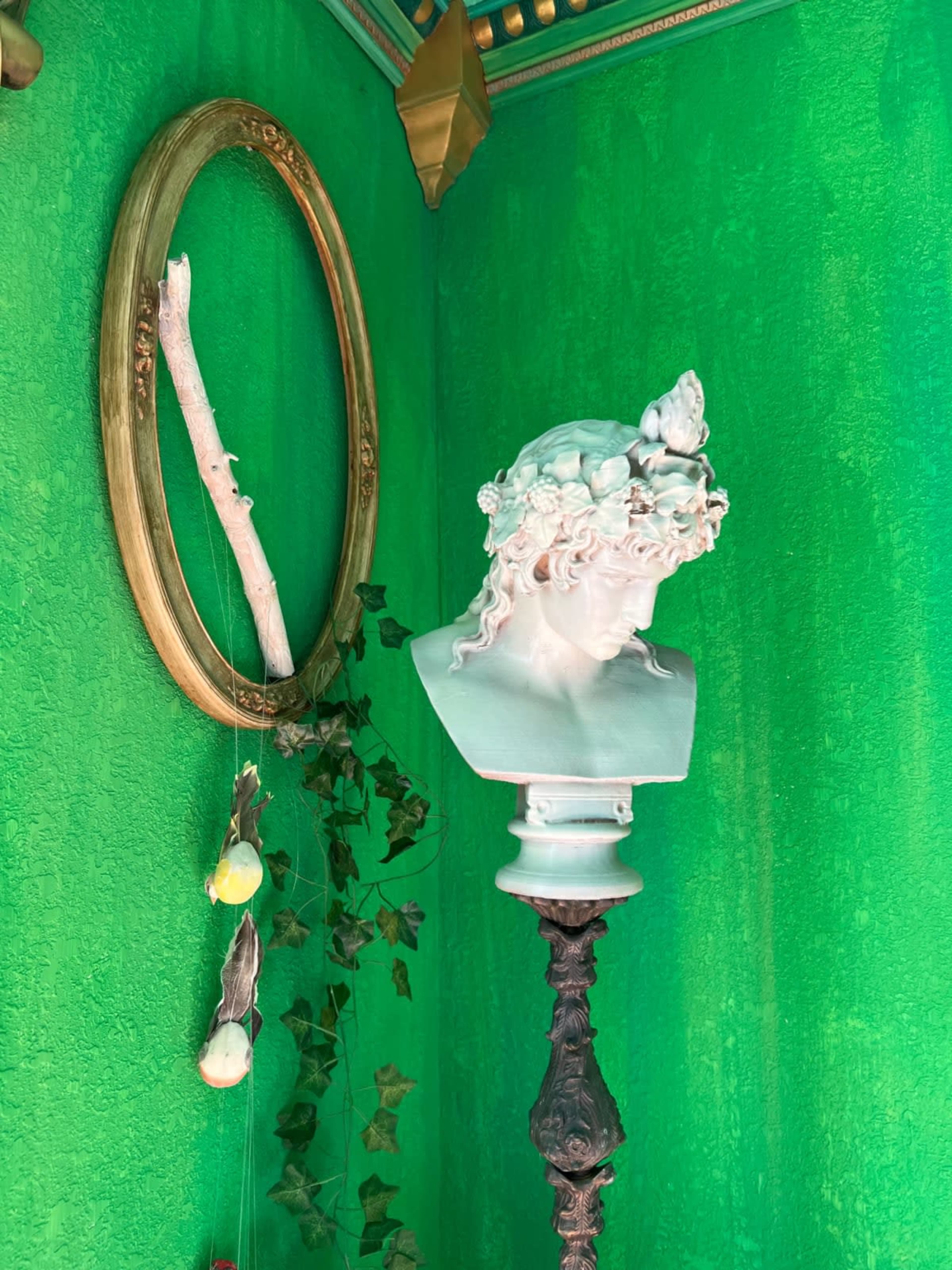 A white bust of a figure is displayed on a dark pedestal against a vibrant green wall, with a circular gold frame and a branch hanging beside it.
