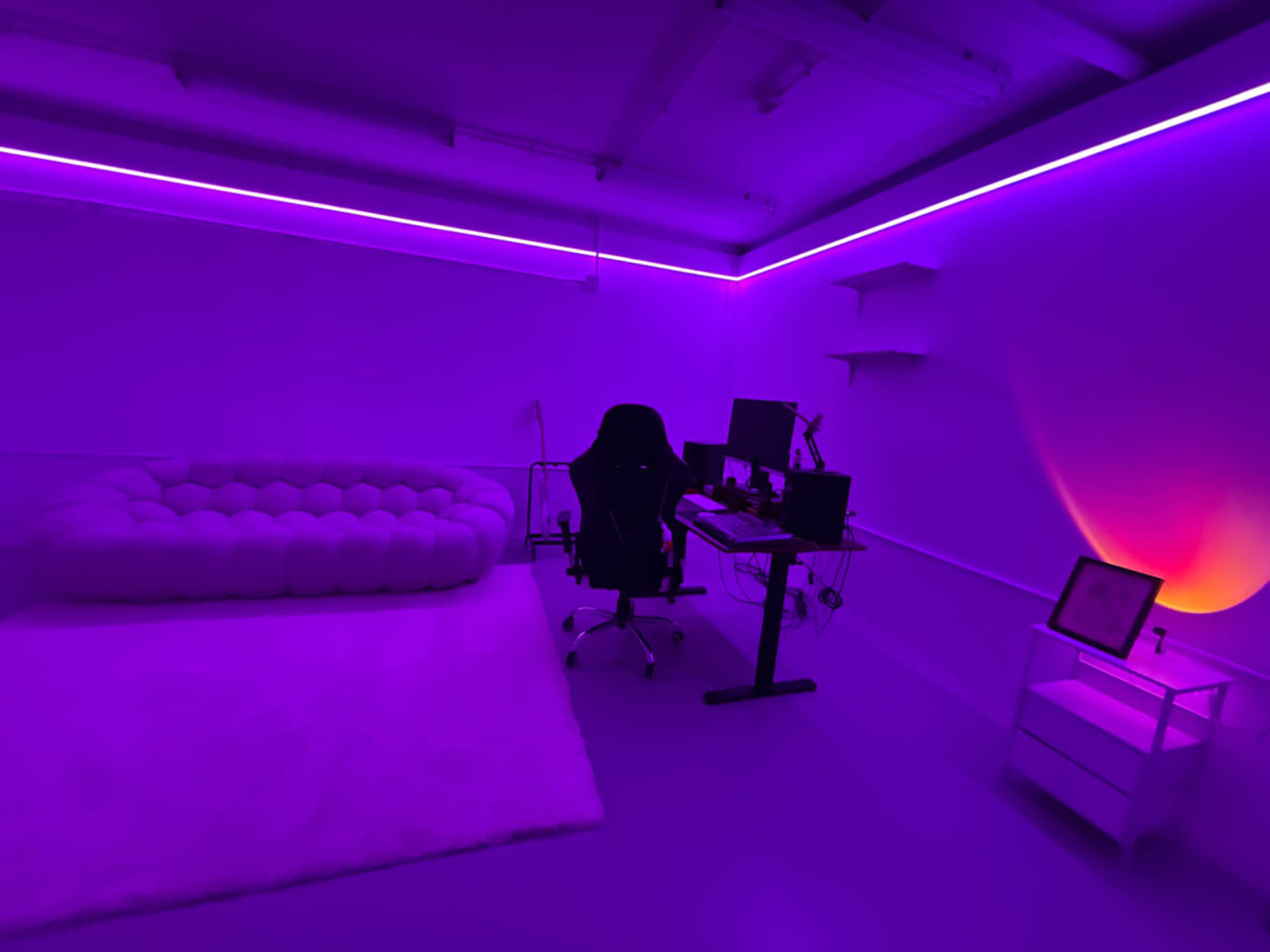 The image shows a modern room with purple lighting, featuring a large plush sofa, an office chair, a desk with two computer monitors, and minimal furnishings.