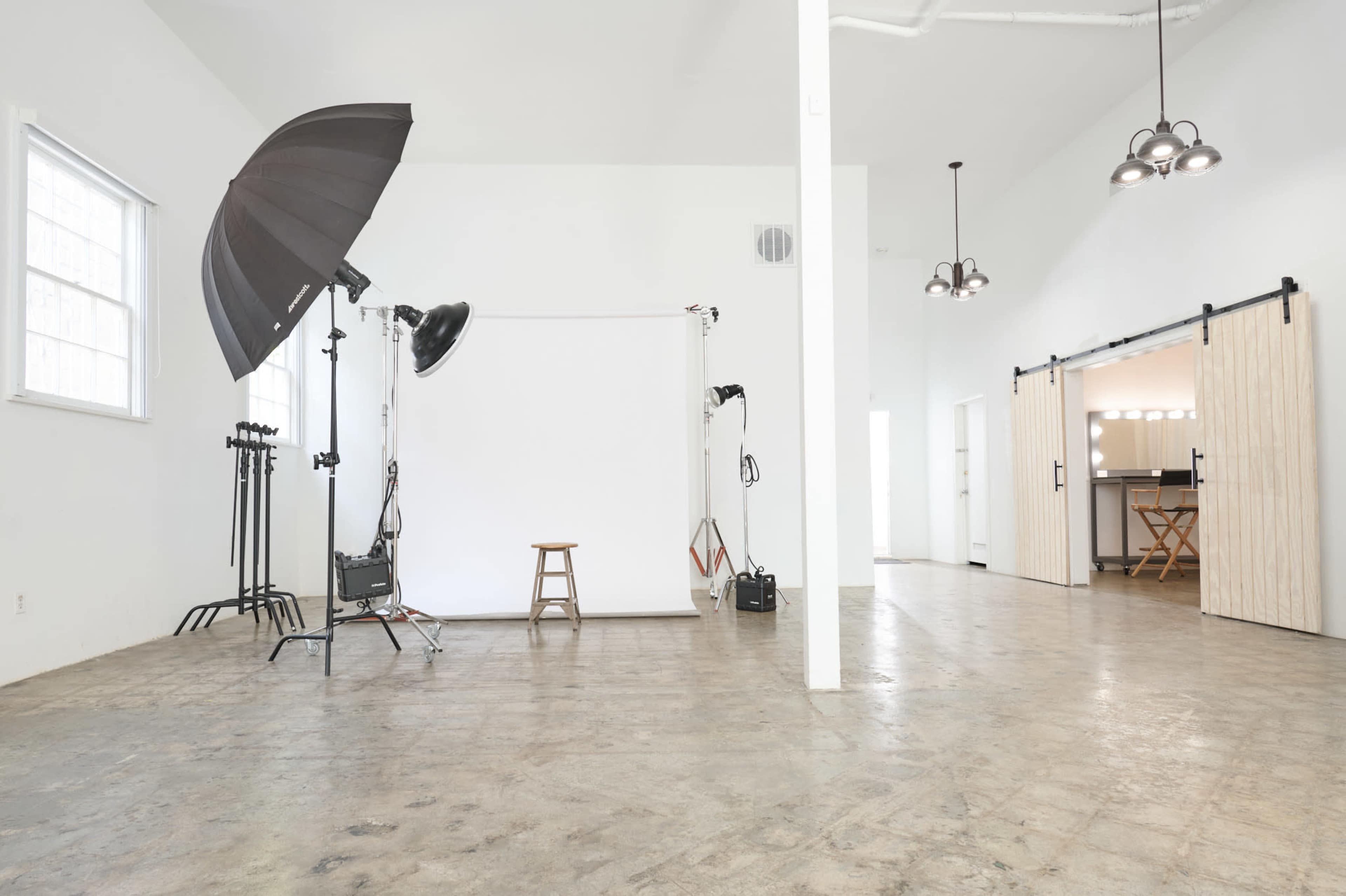 The image shows a spacious photography studio with a plain backdrop, lighting equipment, and minimal furnishings.