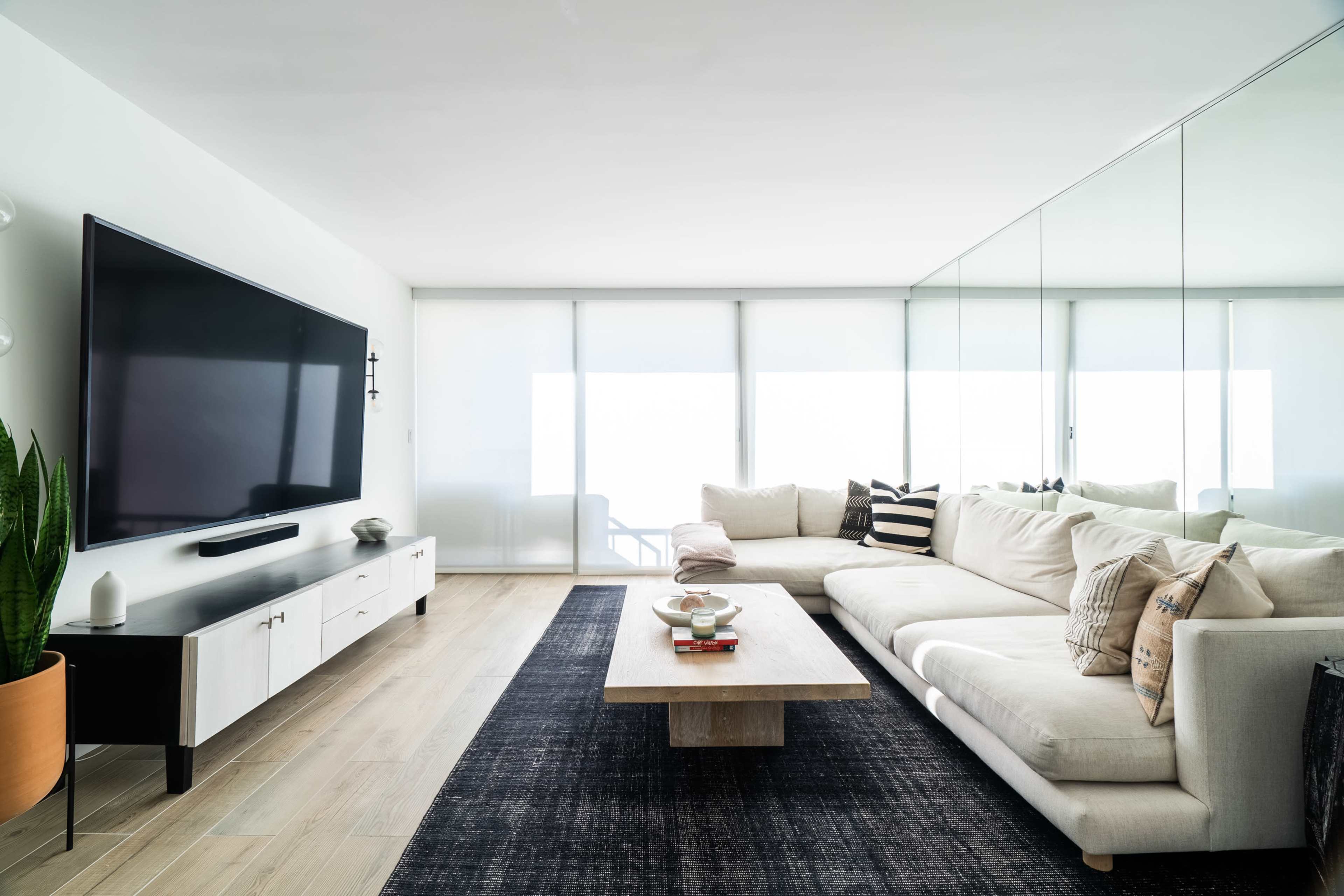 A modern living room with a large sectional sofa, a wooden coffee table, a television mounted on the wall, and large windows covered by sheer curtains.