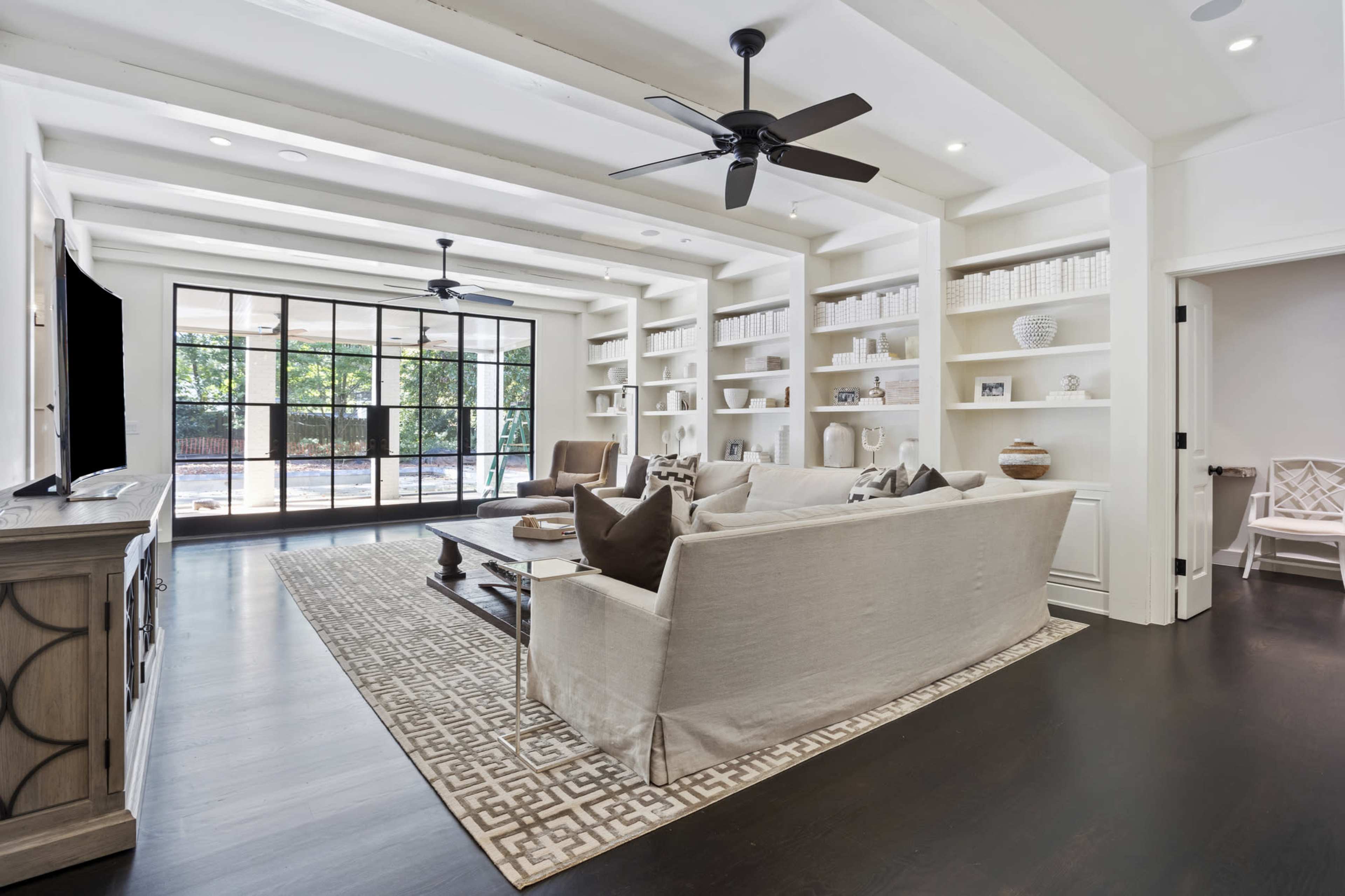 The living room features a light-colored sectional sofa, a large area rug, built-in shelves, and a wall of windows overlooking an outdoor space.