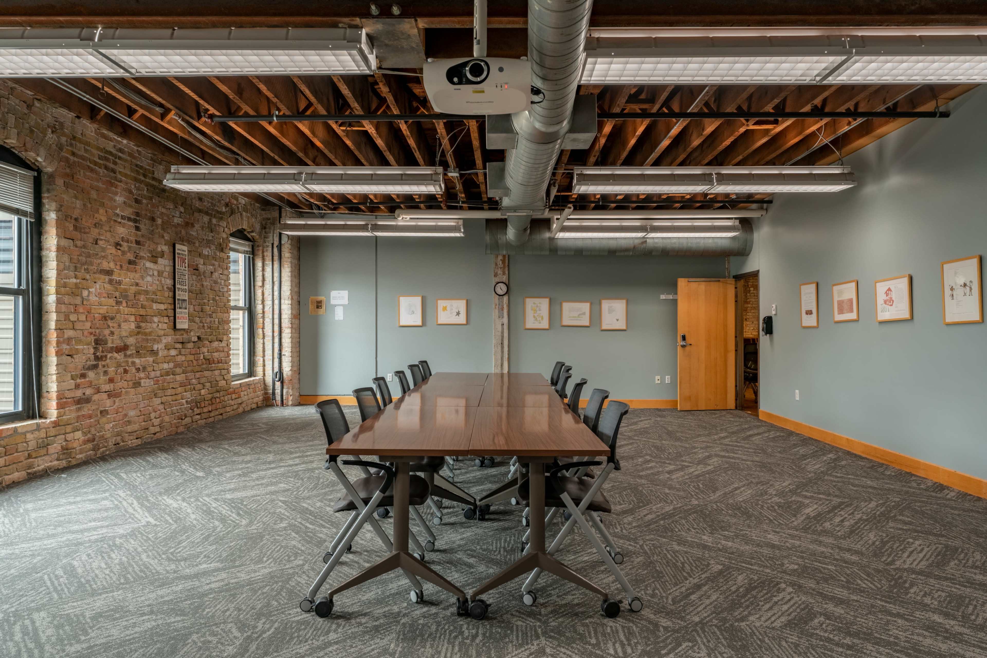 The image shows a spacious conference room featuring a long wooden table with rolling chairs, brick walls, and large windows.