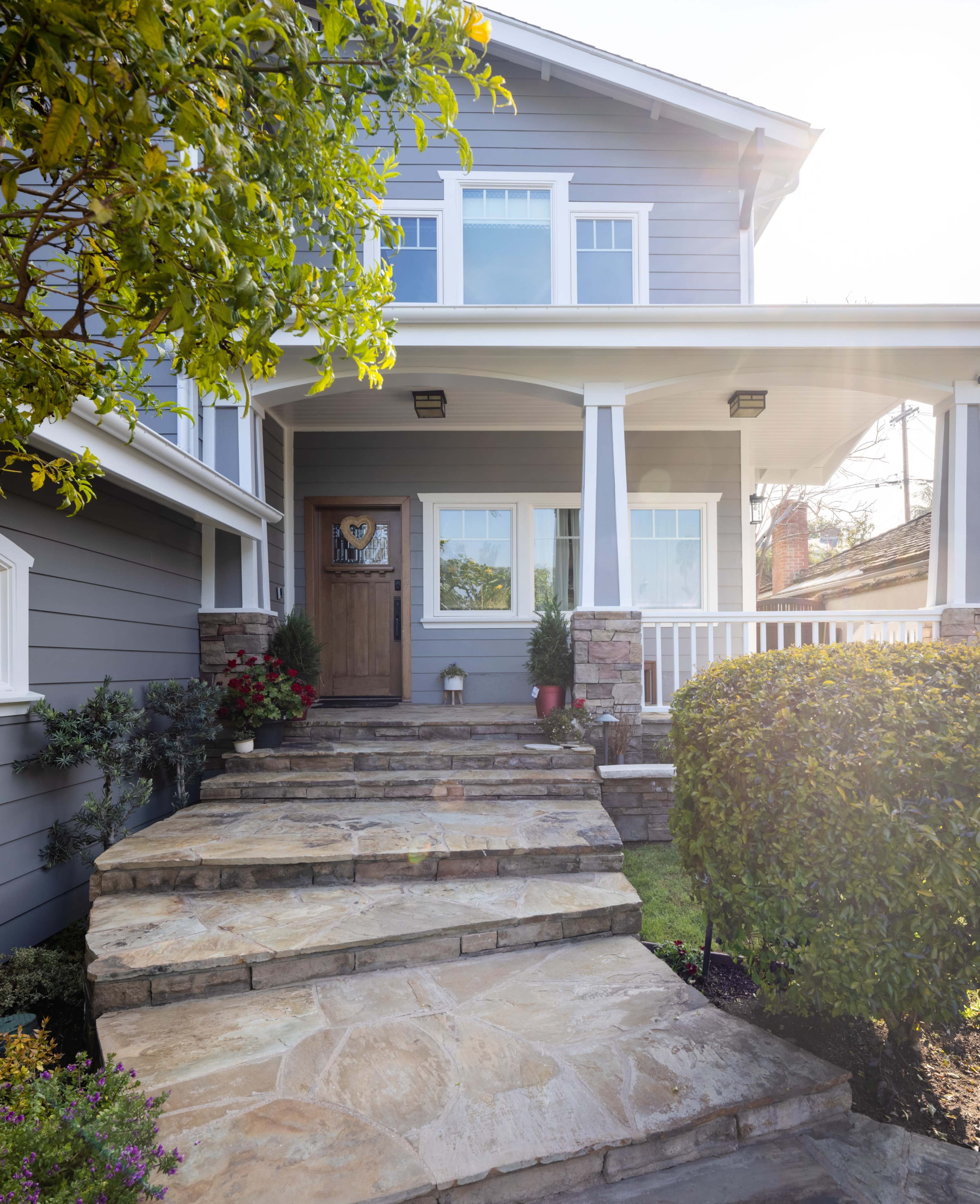 A stone pathway leads to the front door of a gray house with white trim and a porch, surrounded by neatly landscaped shrubs and flowers.