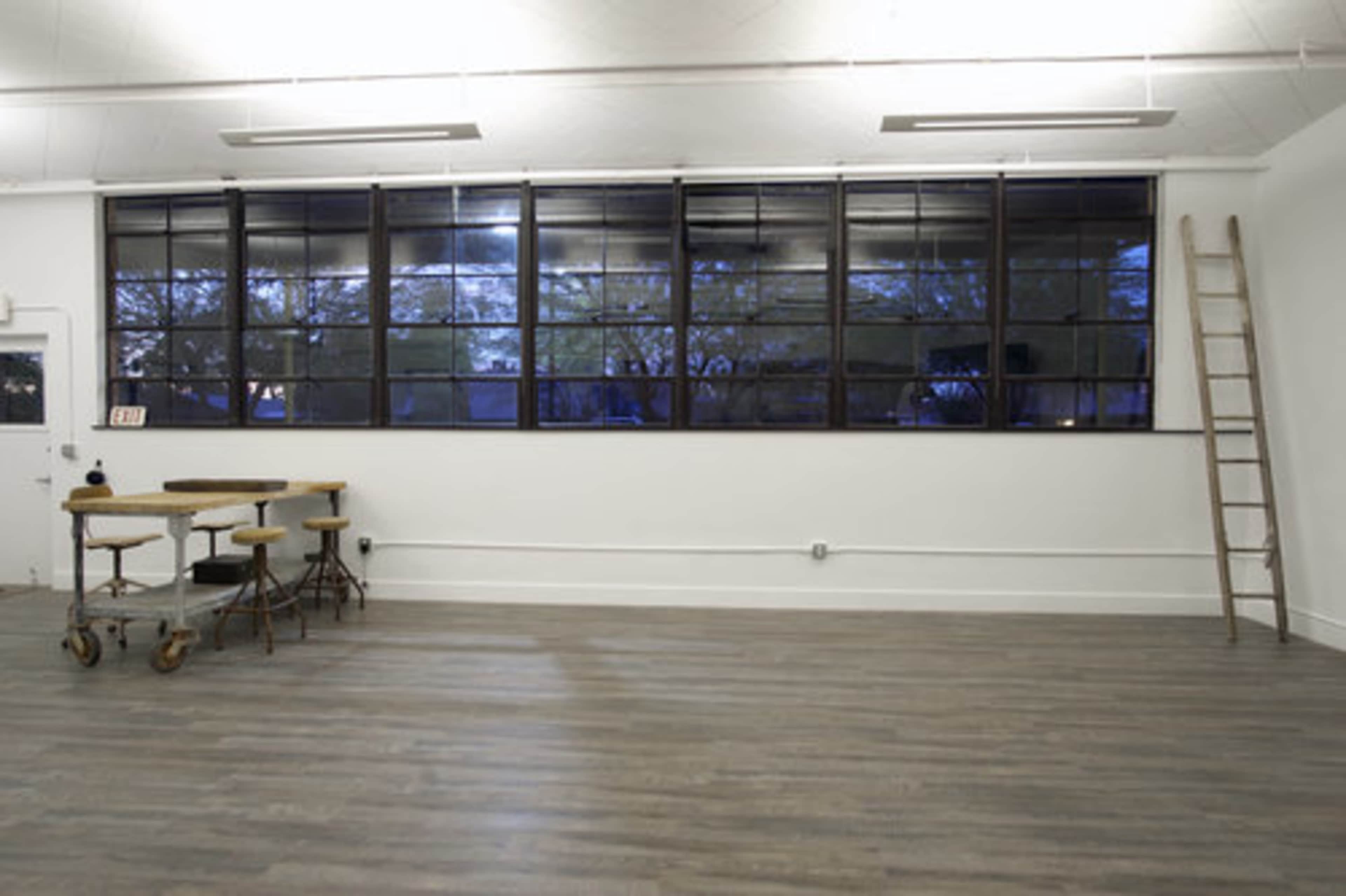 A spacious, well-lit room with large industrial windows, a wooden ladder, and a set of two wooden tables on the floor.