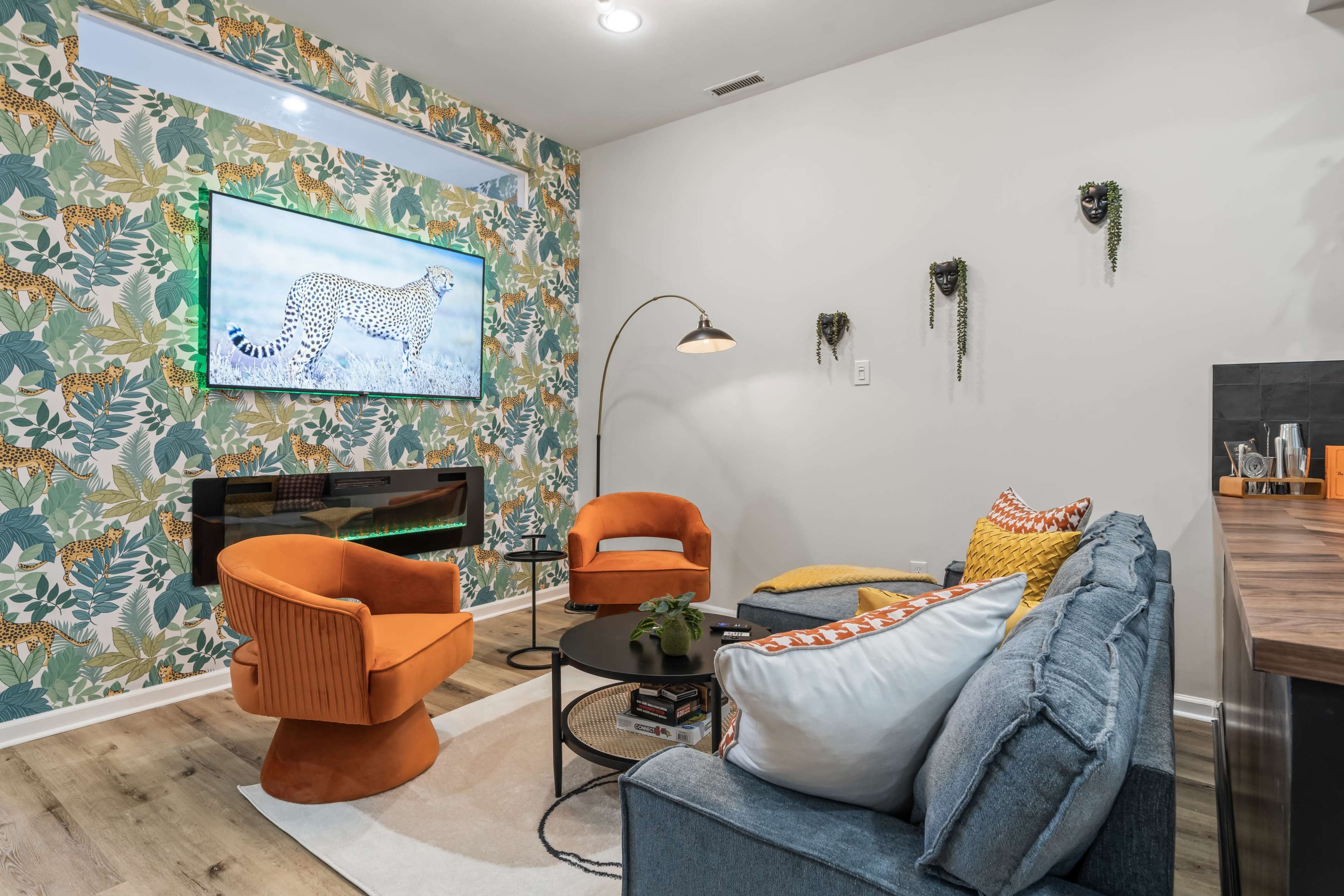 A modern living room features two orange chairs, a blue couch, a wall with vibrant tropical wallpaper, and a wall-mounted television displaying a cheetah.