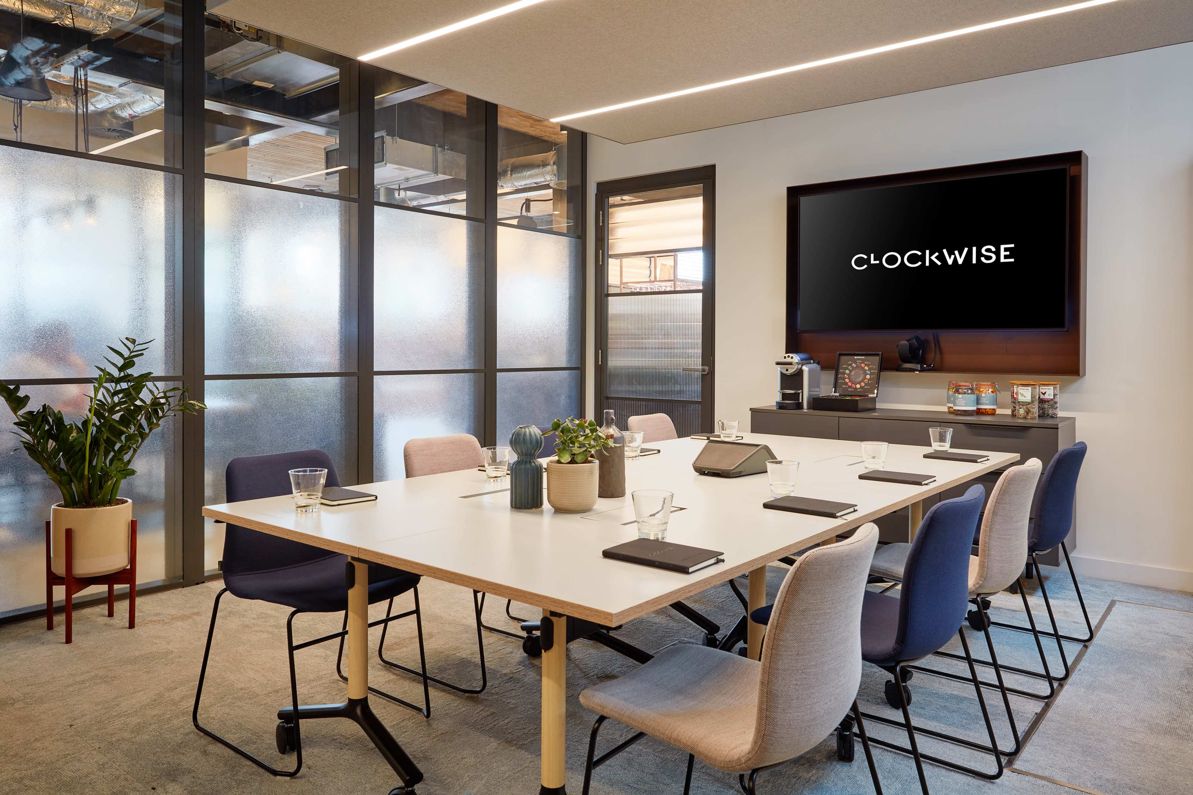 A modern conference room features a large table surrounded by various chairs, a television displaying "CLOCKWISE," and glass walls for visibility.
