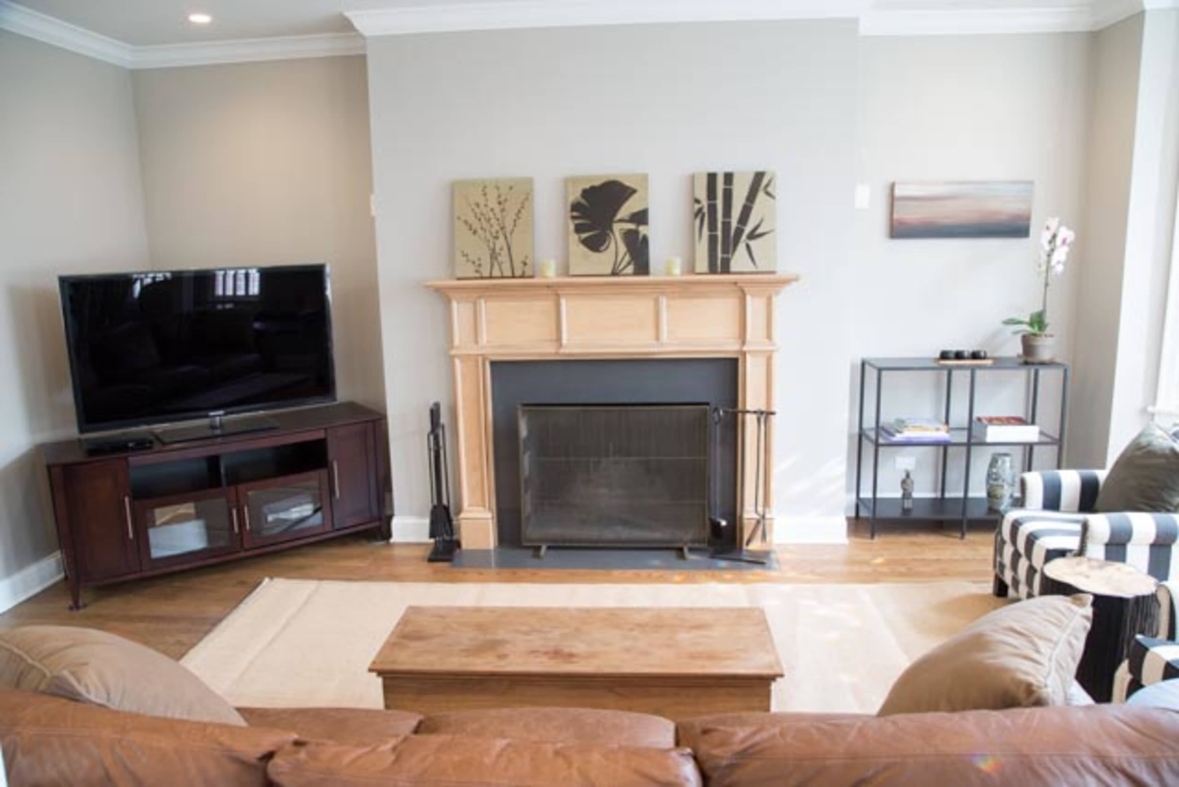The living room features a brown leather sofa, a wooden coffee table, a large television, and a fireplace flanked by decorative artwork.