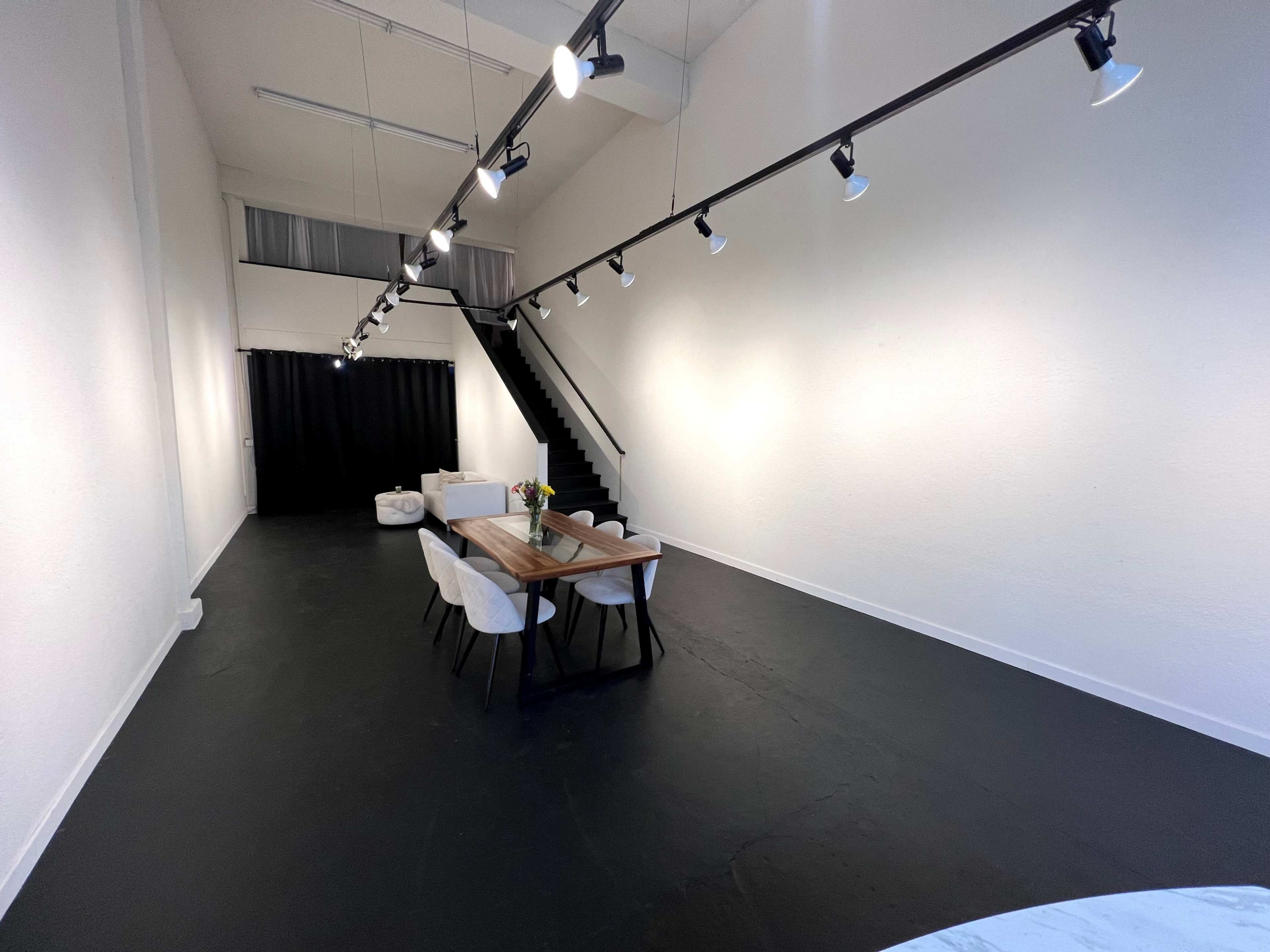 A spacious, brightly lit room with a wooden dining table surrounded by white chairs, a staircase in the background, and a black curtain to the left.