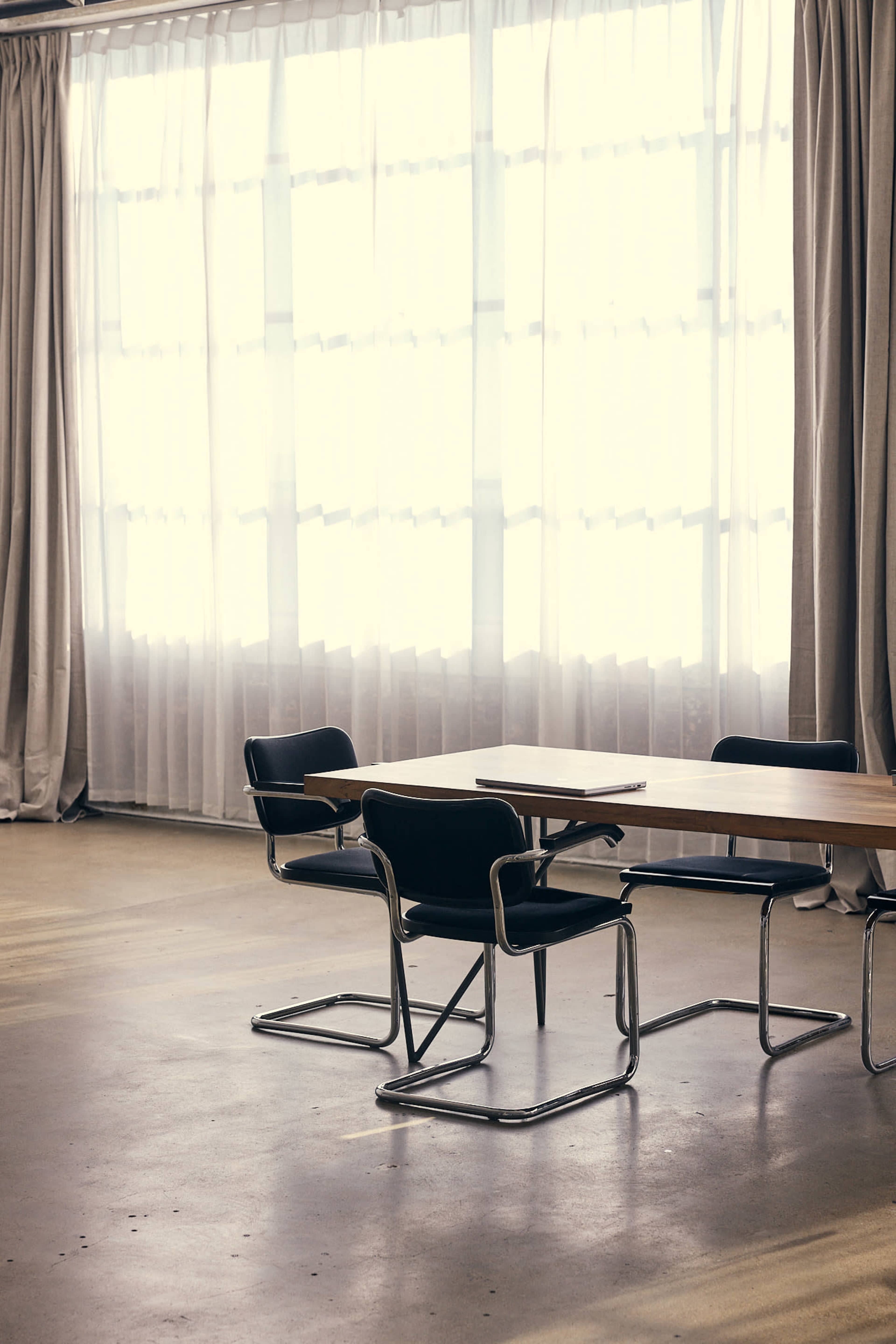 A simple meeting room features a wooden table with three black chairs set against large windows covered with sheer curtains.