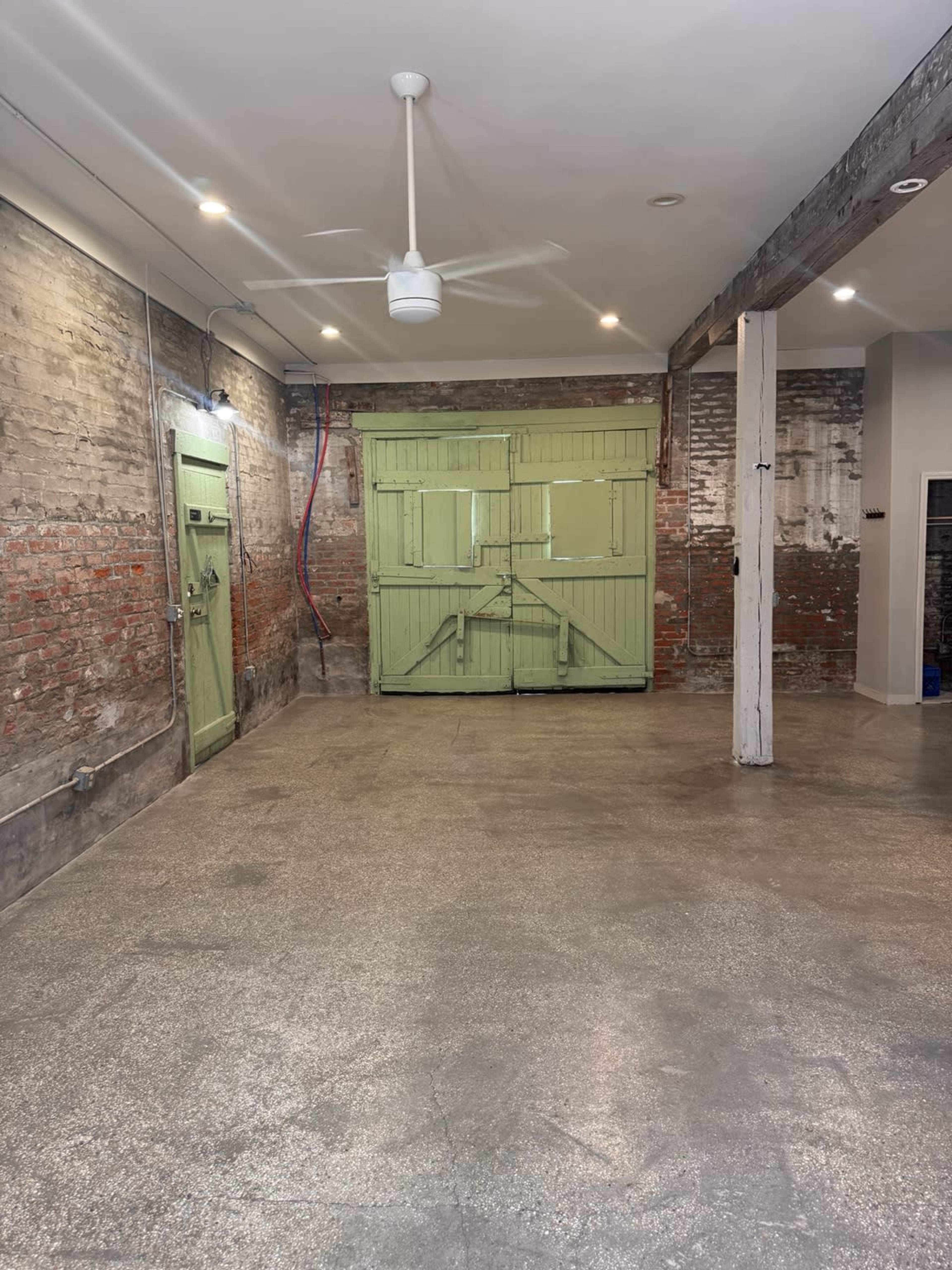 The image shows an empty room with exposed brick walls, a green double door, and a concrete floor, illuminated by ceiling lights.