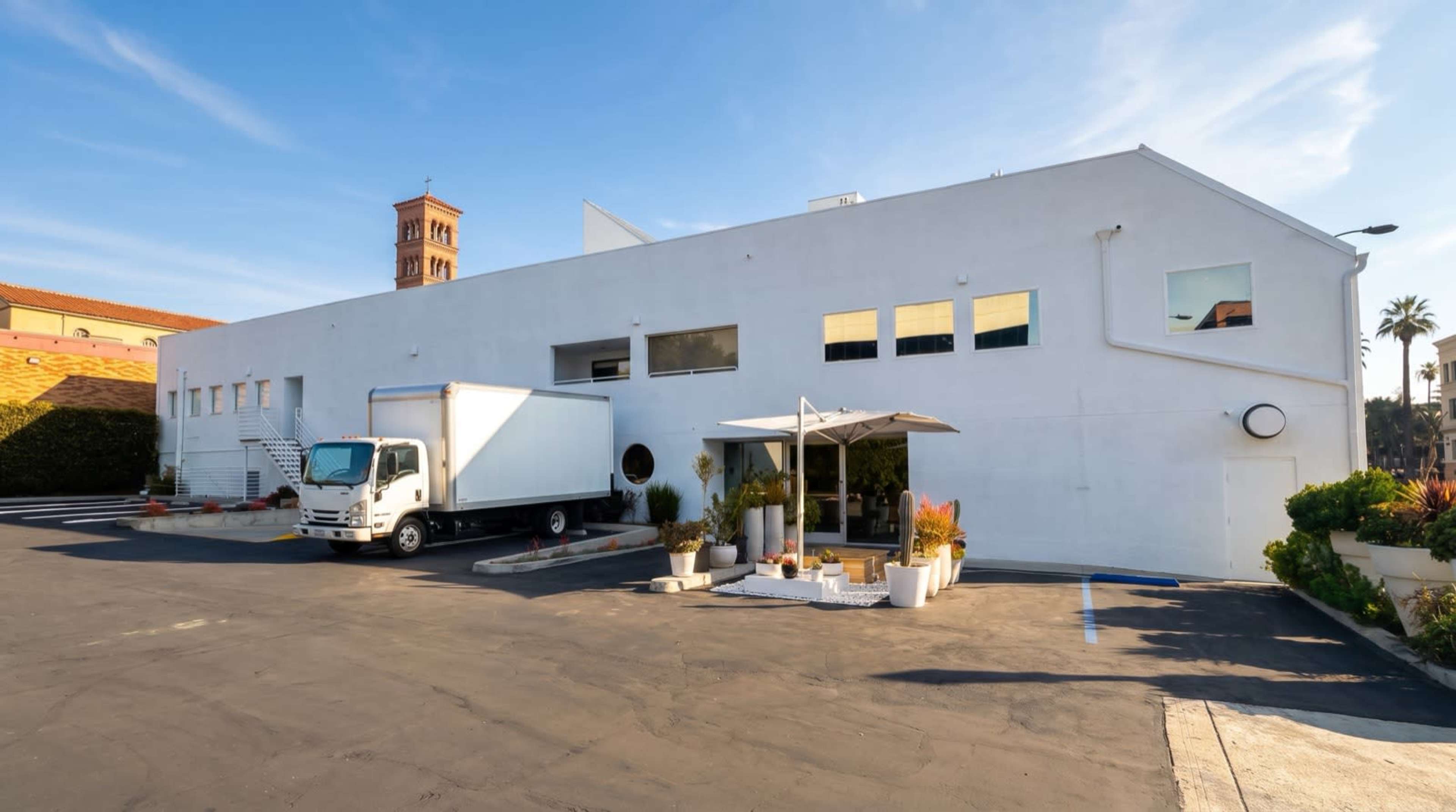 A white building with multiple windows and a loading truck parked in front is situated in a parking lot.