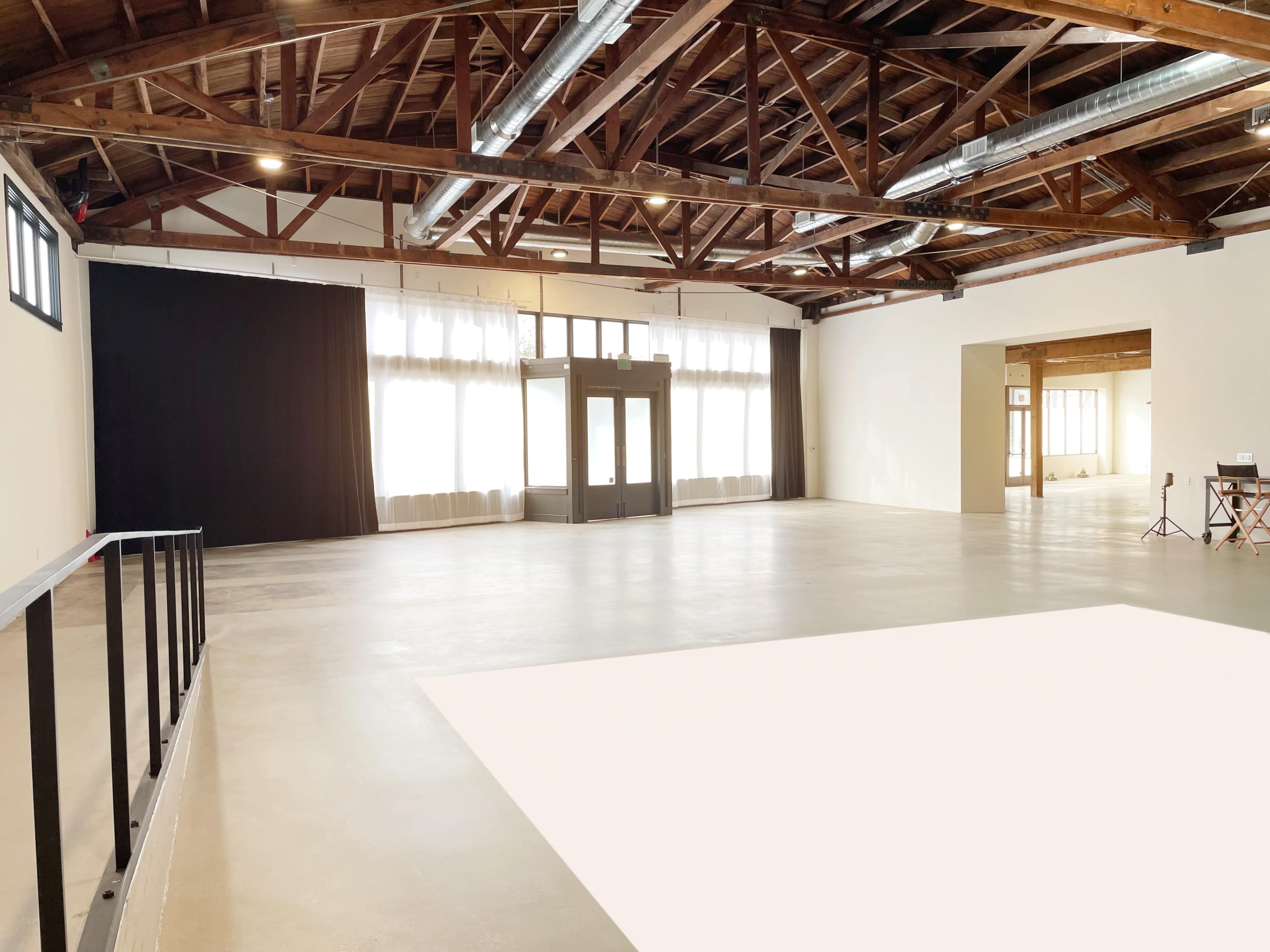 A spacious, well-lit interior of a warehouse with wooden beams, large windows, and a black curtain at one end.